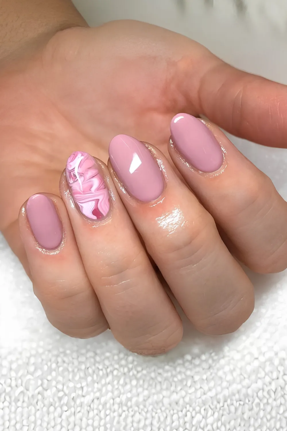 A detailed photograph of a hand resting on a white textured towel, showcasing an elegant mauve-pink manicure on short, oval fingernails. The index, middle, pinky, and thumb feature a solid, glossy dusty rose polish, while the ring finger serves as a marble accent nail with swirling veins of pink, white, and deep magenta.