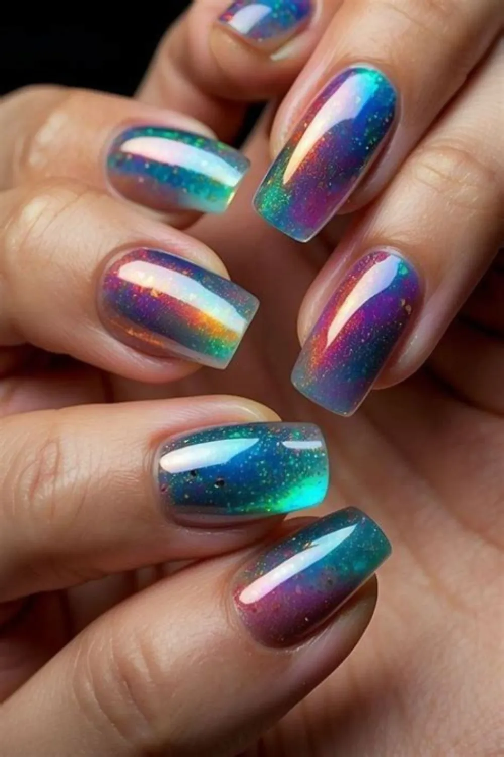 A close-up photograph showcasing a breathtaking galaxy aurora manicure on long, square-cut nails. The nail art features a dark, translucent base filled with fine iridescent glitter and a magnetic cat-eye shimmer overlay that shifts dramatically between deep teal blue, violet, magenta, and shimmering golden-orange depending on the light angle.