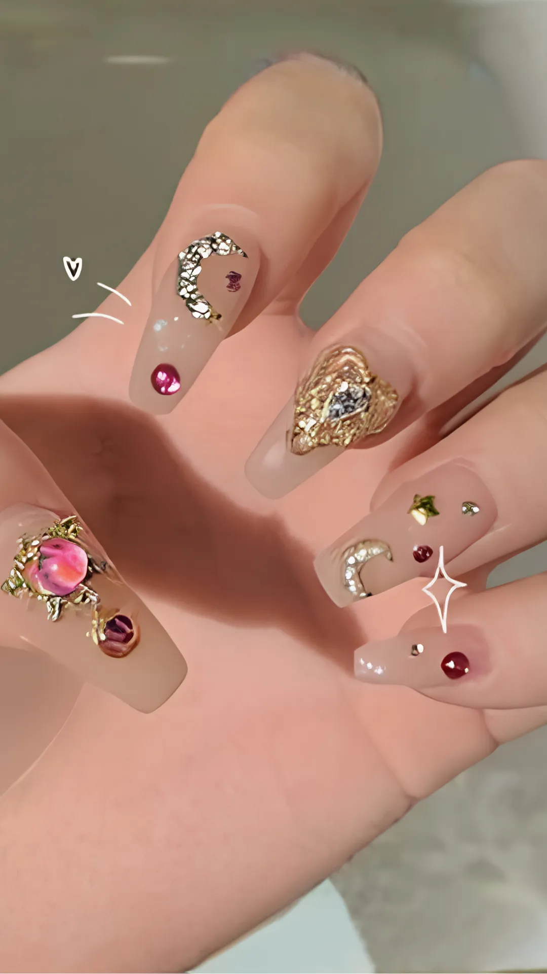 A close-up photograph of long, square-shaped acrylic nails featuring an ornate, celestial-themed Korean nail design. Against a glossy, sheer nude base, each nail is elaborately decorated with heavy 3D charms, including chunky gold metallic crescent moons, a large textured golden heart embedded with a central rhinestone, pink gem studs, and small gold stars.