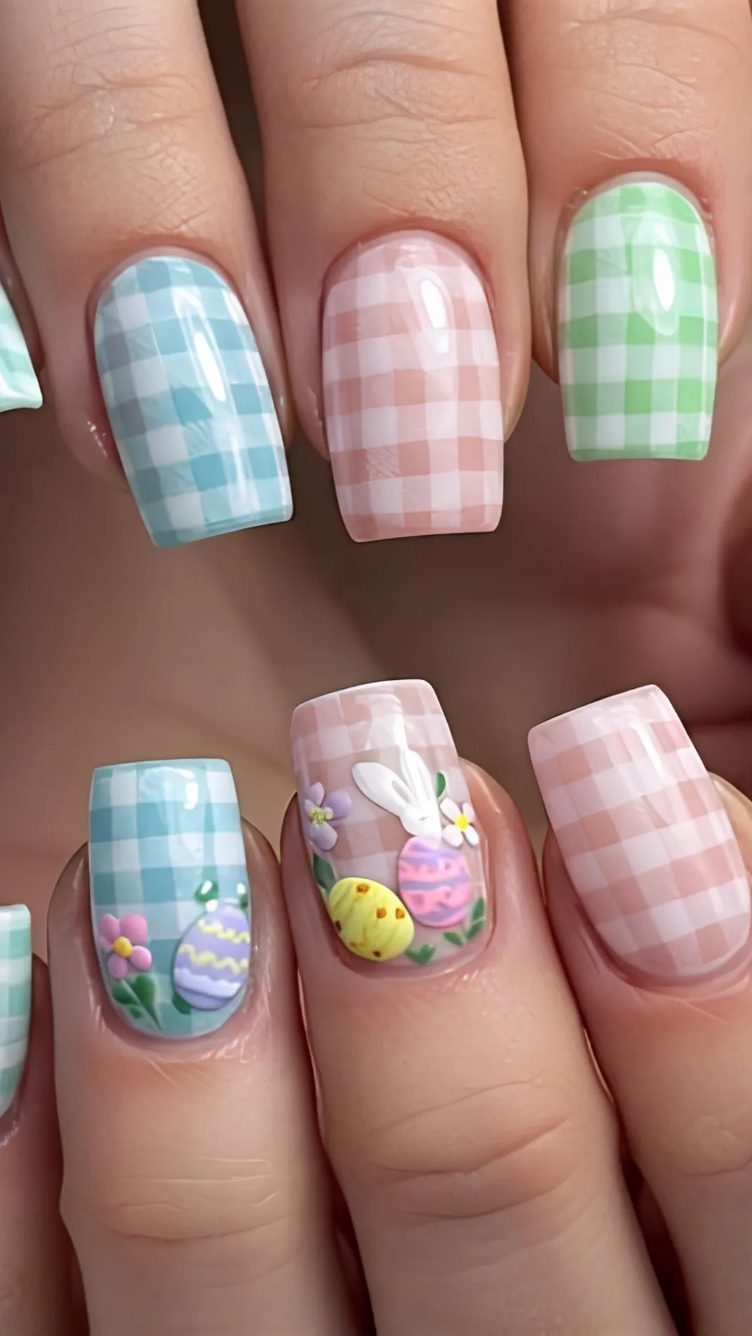 A detailed close-up of a charming pastel gingham or plaid manicure on square-shaped nails. The fingernails feature alternating pastel grid patterns in sky blue, soft baby pink, and light sage green over a white base. The accent nails are decorated with embossed, 3D textured elements including a white Easter bunny hiding behind colorful decorated eggs, small purple and white spring flowers, and delicate green leaves.