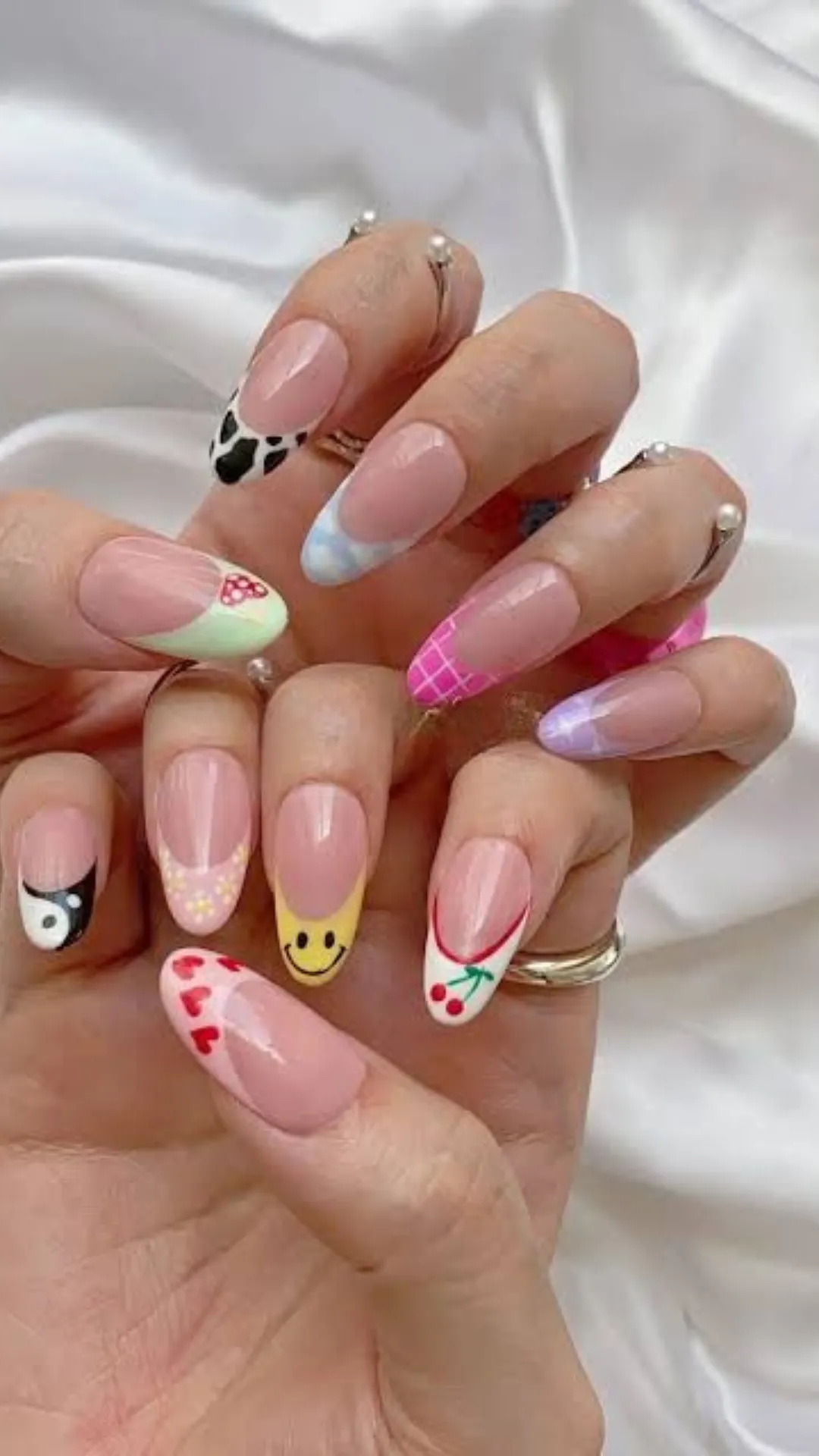 A detailed photograph focuses on both hands displaying a dynamic "Skittles" style mix-and-match nail art on short almond-shaped nails. Against a sheer nude base, each nail features a unique design over distinct colored tips or patterns: lime green with a cherry decal, bright yellow with a black smiley face, pink checkerboard, purple with abstract waves, sky blue with clouds, red with hearts, abstract cow print, yin-yang symbol, and pink with a single green dot. The diverse, cheerful patterns create a joyful, optimistic look that remains modern and elegant on short nails.