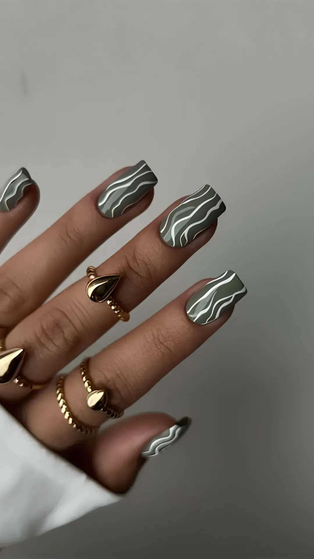 Modern square nails coated in a muted matte olive-grey polish, detailed with wavy, organic abstract lines in white and light sage green.