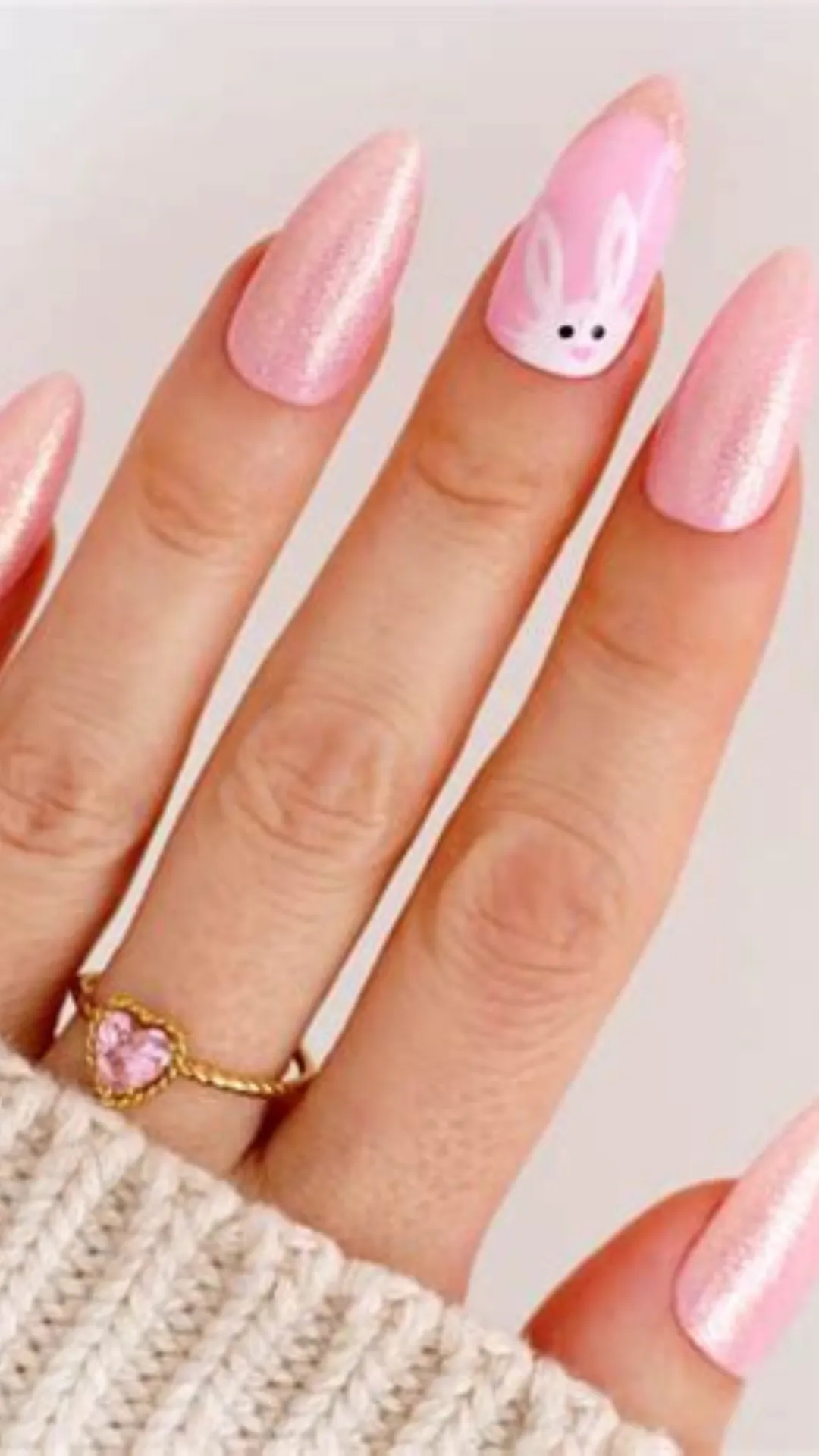 A close-up shot of a hand wearing a delicate gold ring with a heart-shaped pink gemstone, showcasing a shimmering spring manicure. The long, almond-shaped nails are coated in a pale pink polish filled with fine, iridescent glitter that gives a subtle holographic glow. The ring finger features a cute accent design with a hand-painted white Easter bunny face looking upward from the cuticle area.