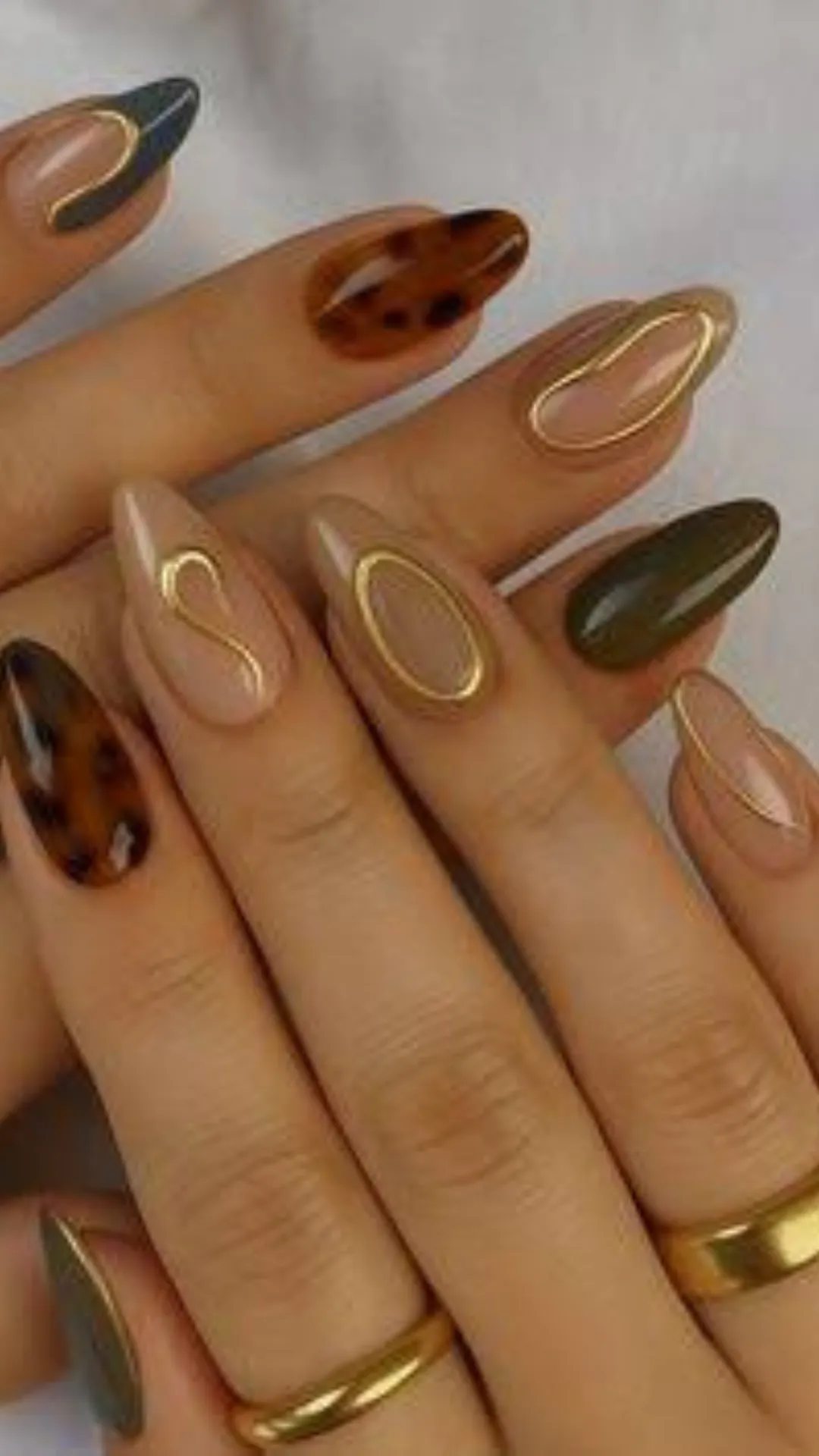 A detailed close-up of a pair of hands showcasing a sophisticated, multi-textured autumn-inspired manicure on long, oval-shaped nails. The eclectic mix-and-match design features translucent amber tortoiseshell patterns with dark brown splotches, solid glossy olive-green nails, and various nude-colored bases. The nude nails are elegantly accented with raised, 3D metallic gold gel outlines formed into abstract wavy frames and organic circular borders. The model is also wearing thick, polished gold bands on their fingers, complementing the metallic elements of the nail art.
