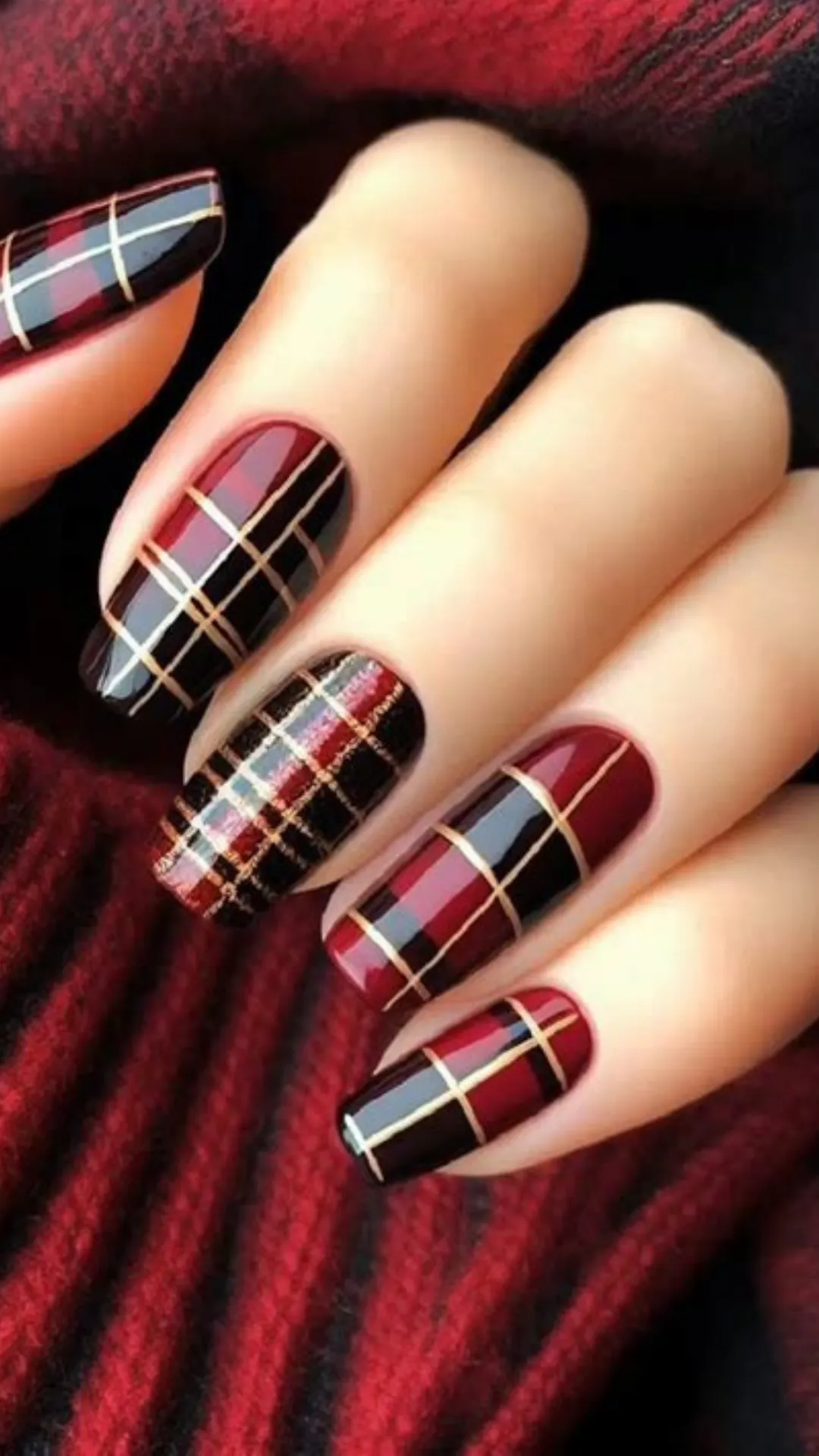 Cozy autumn and winter nail design showcasing long oval nails painted in a glossy red, black, and gold metallic plaid pattern, paired against a red cable-knit sweater.