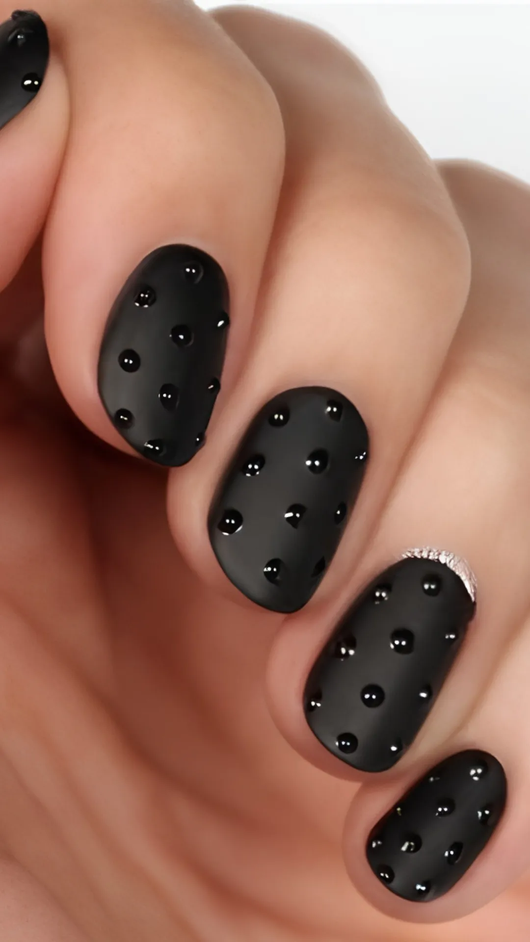 A close-up of a dark and textured minimalist manicure featuring short round nails in a matte black finish, detailed with glossy black raised 3D water droplets and a tiny silver glitter accent at the cuticle line.