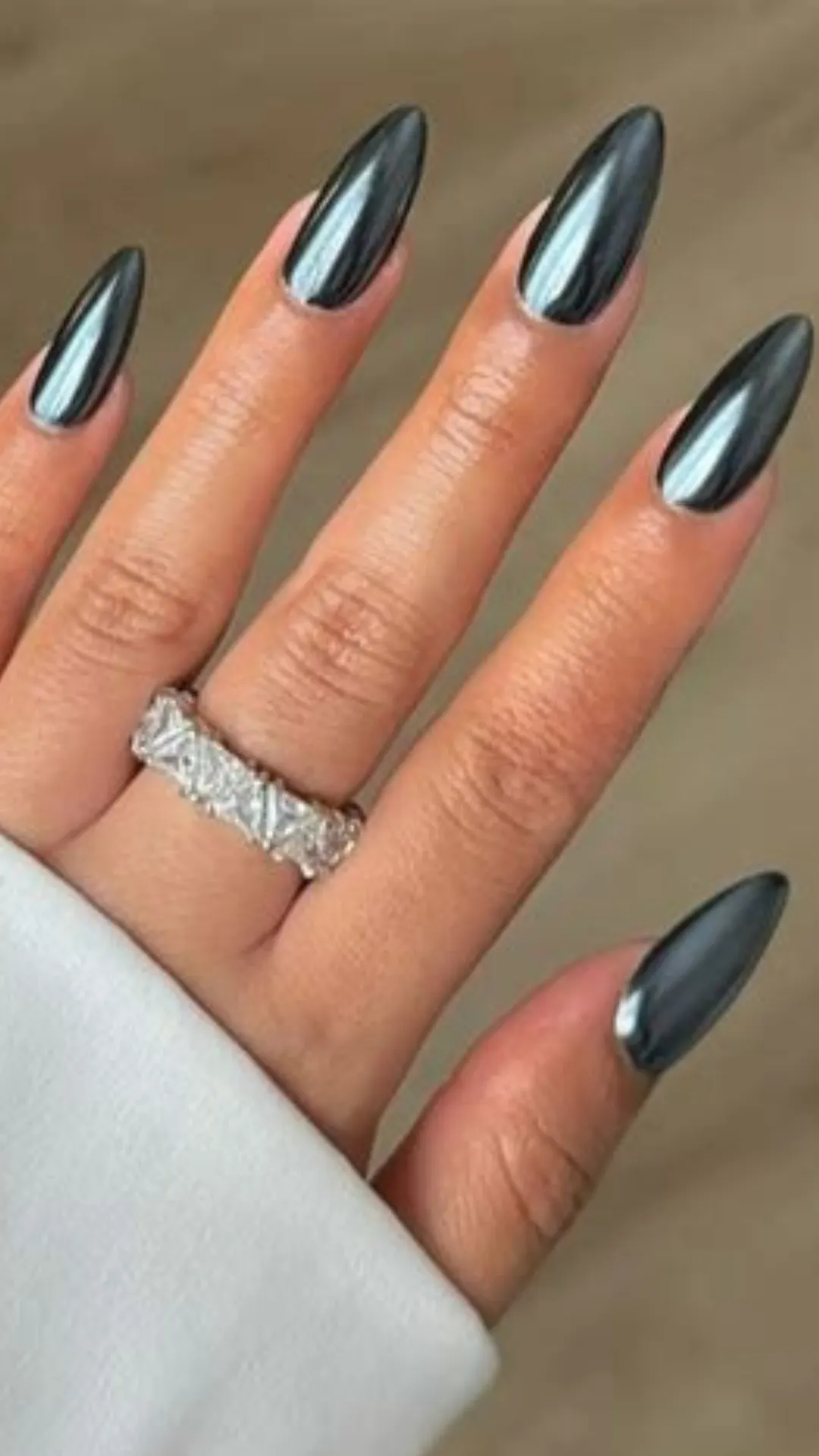 A detailed close-up of a hand featuring a striking, futuristic dark gunmetal chrome manicure on long, sharp stiletto-shaped nails. The highly reflective metallic finish has a mirror-like shine that shifts between silver and deep charcoal tones under the light. A thick, sparkling silver ring covered in emerald-cut cubic zirconia or diamonds is worn on the ring finger, adding an extra layer of luxury.