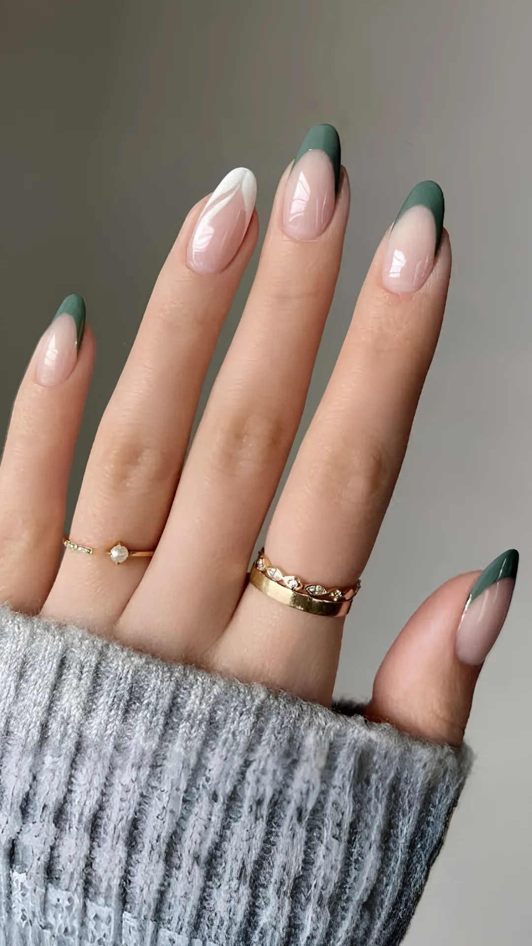 A close-up view focuses on a left hand featuring modern French tip almond-shaped nails. Four nails display a deep forest green tip, while the middle finger showcases a crisp white tip, all applied over a sheer nude base. The hand wears several gold rings, including a simple band and one set with small diamonds. The hand is positioned near the ribbed cuff of a grey cable-knit sweater, adding a soft, cozy texture. The elegant everyday look is achieved through the mix of classic French tips and a sophisticated, trendy color palette.
