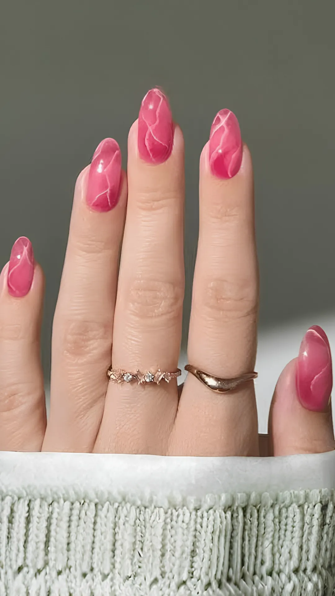 A detailed close-up shot of a hand displaying long, almond-shaped nails with a sophisticated rose quartz marble effect. The nails feature a rich, translucent berry-pink base interwoven with realistic, fine white veining that mimics polished gemstone crystals. The hand, wearing two delicate gold accent rings with small embedded gems, rests against the textured cuff of a light green knit sweater.