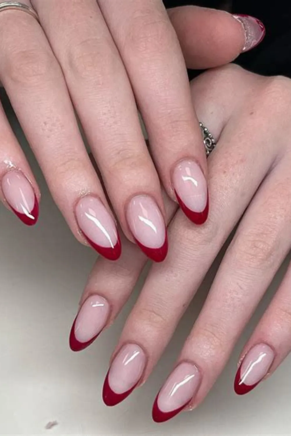A detailed close-up shot of two hands displaying an elegant and timeless interpretation of a French manicure. The long, almond-shaped fingernails are styled with a glossy, semi-translucent pale pink base coat. Instead of traditional white, the tips are painted with clean, perfectly curved smiles in a deep, sophisticated cherry red lacquer.