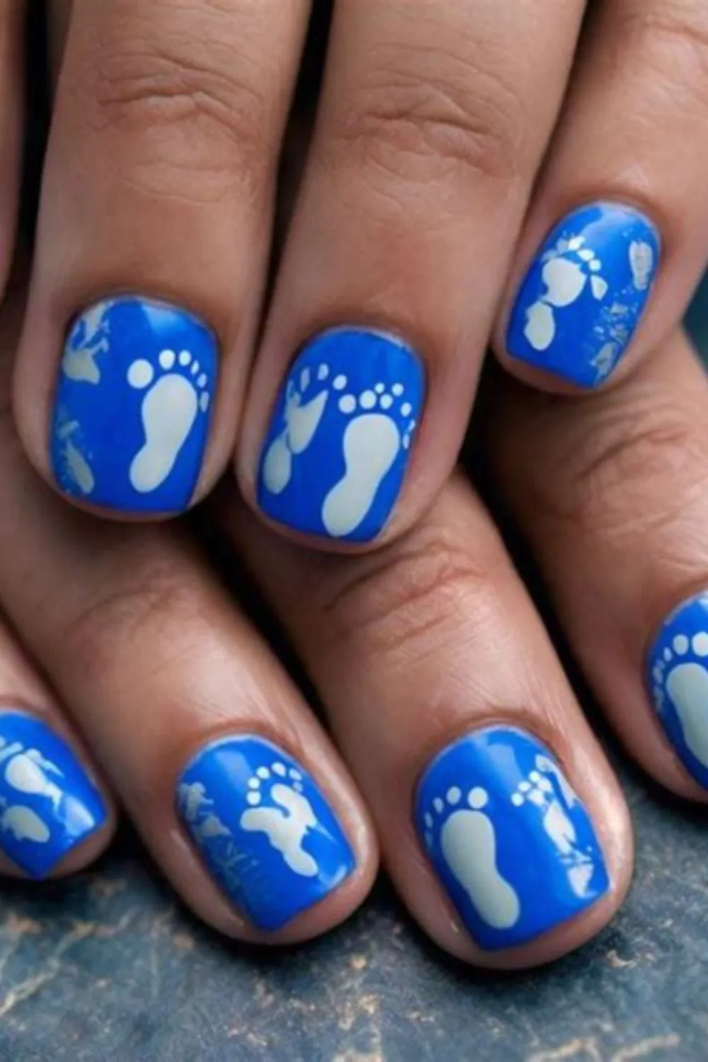 An extreme macro photograph showing a royal blue baby boy celebration manicure on short, rounded fingernails. The nails are painted in a rich, full-coverage glossy royal blue polish. Each nail bed is stamped or hand-painted with crisp white miniature baby footprints and small handprints, creating a bold, joyful, and unambiguous gender-reveal statement.