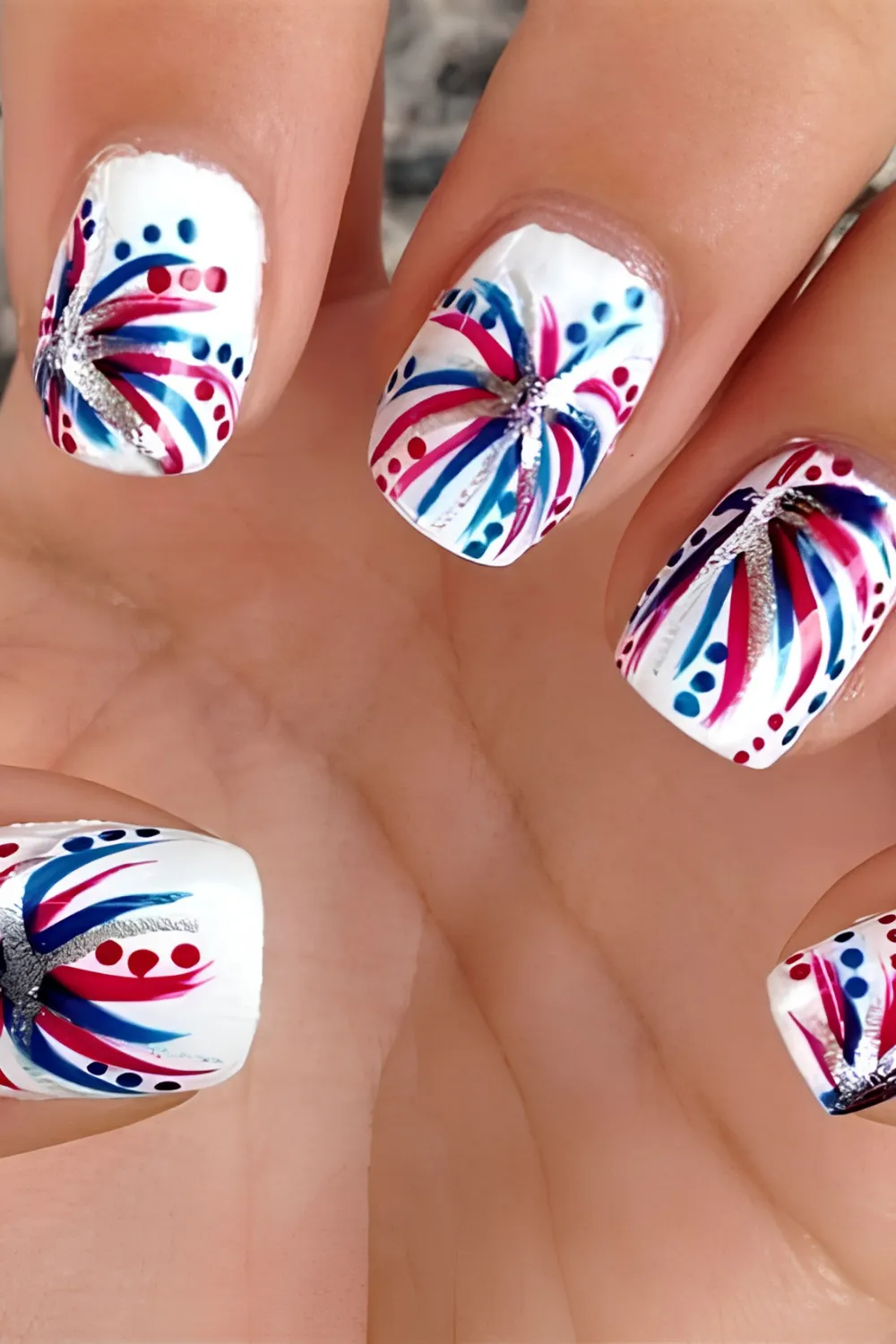 An extreme macro photograph showcasing a dynamic firework-themed holiday manicure on short, squared nails. Each nail features a crisp, opaque white background decorated with vibrant, exploding firework bursts made of curved red, royal blue, and metallic silver glitter streaks, accented by tiny surrounding red and blue polka dots.