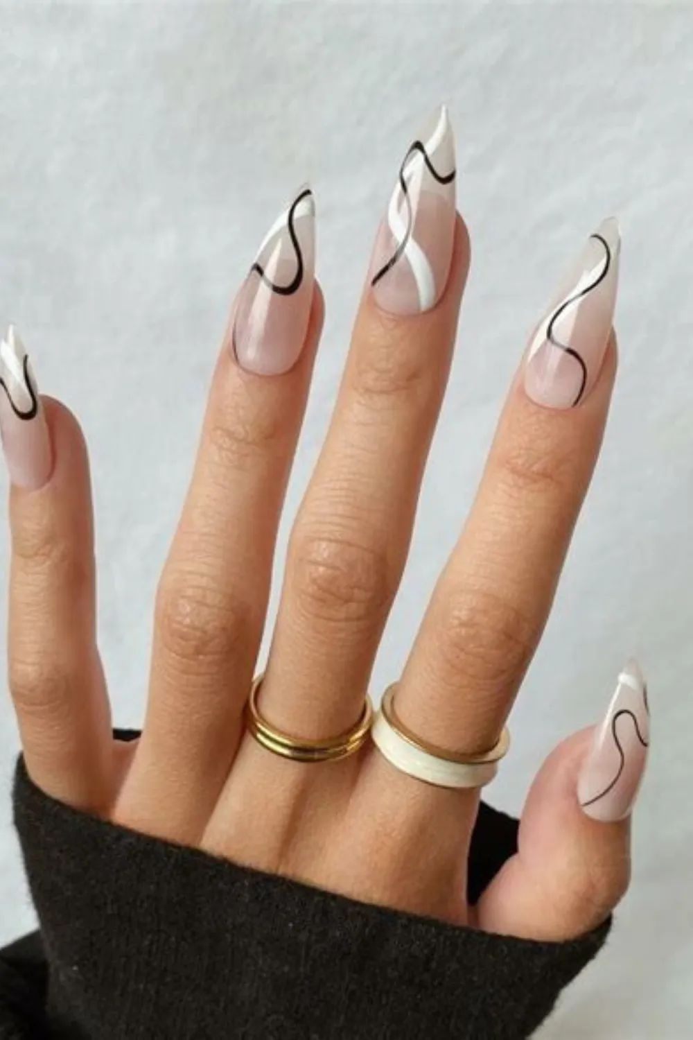 A detailed photograph of a hand displaying long, stiletto-shaped nails with a modern abstract design. Set against a sheer, high-gloss nude base, each nail features flowing, hand-painted wavy lines in solid white and stark black that curve along the edges, paired with delicate gold accent rings and a white enamel band worn on the fingers.