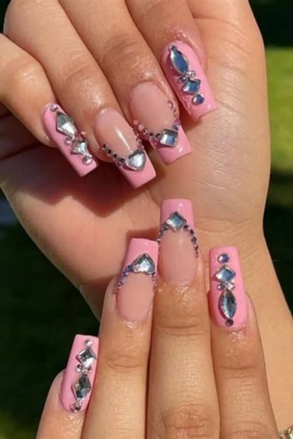 A detailed close-up shot of two hands displaying a maximalist Y2K rhinestone manicure on long, coffin-shaped nails. The nails are painted in a bubblegum pink gel, featuring classic French tip outlines lined with sparkling silver crystals. Every nail is heavily embellished with large, multi-shaped rhinestones including diamonds, hearts, and marquise gems.