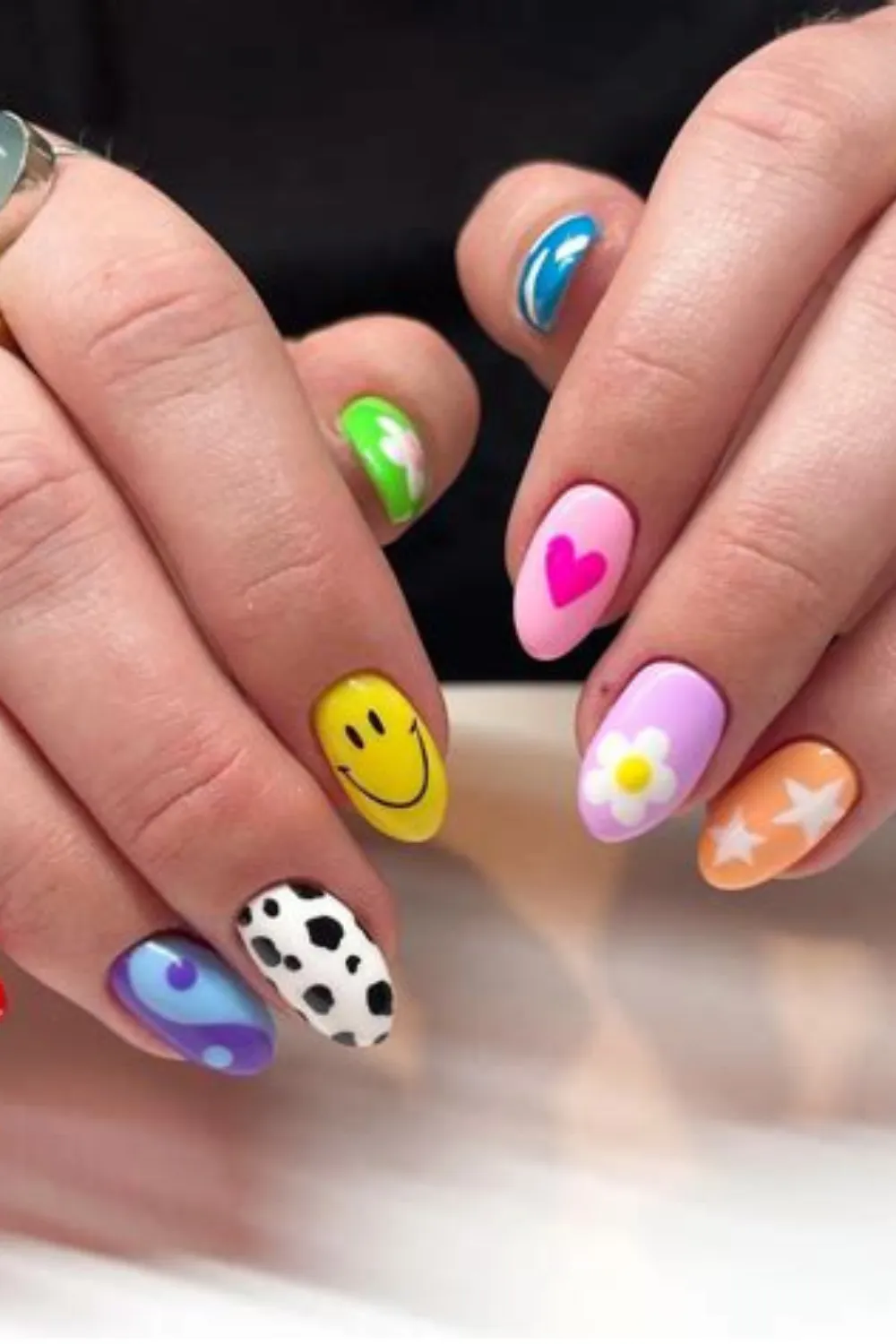 A detailed photograph of two hands featuring a playful, pop-art maximalist gel manicure on short, oval-shaped nails. Every fingernail presents a unique pastel base decorated with fun graphic art, including a bright yellow smiley face, a hot pink heart, a white daisy, white stars over an orange base, a black-and-white cow print, and a purple yin-yang symbol.
