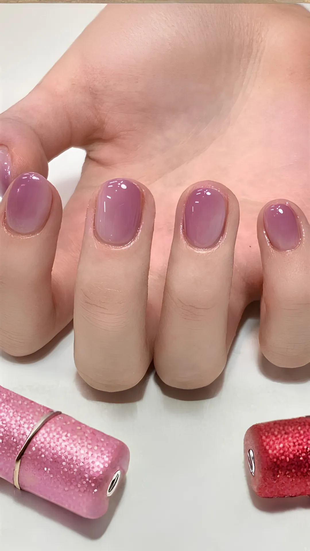 Close-up of a hand displaying a minimal yet elegant Korean gel manicure. The short, rounded nails are coated in a glossy, translucent mauve-pink jelly polish that creates a soft, glass-like aesthetic. The background is a clean white surface featuring two sparkling, glitter-textured lipstick tubes or cosmetic items in shades of bright pink and deep ruby red.
