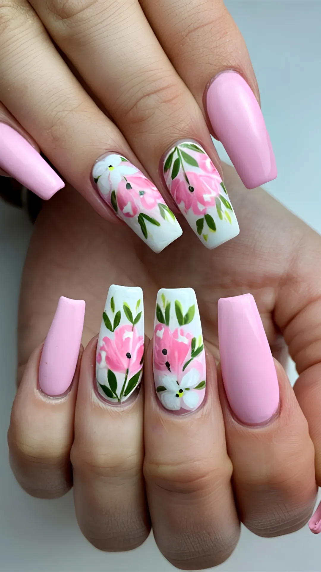 A detailed close-up showing a manicure with long, square-tipped coffin nails perfect for a spring vibe. The thumb, index, and pinky fingers feature a smooth, glossy pastel pink polish. The middle and ring fingernails stand out with a crisp white base decorated with beautifully painted, vibrant pink flowers and trailing deep green leaves, creating a fresh, botanical look.