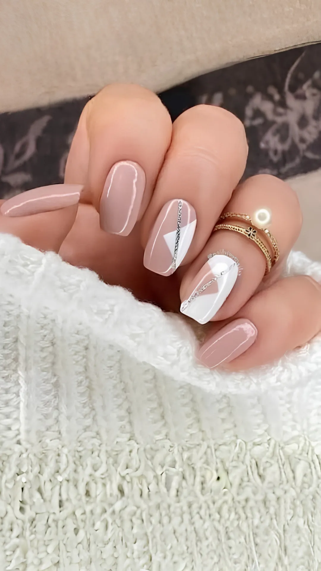 A close-up photograph of a hand showcasing a modern geometric manicure on short, squared nails. The design utilizes a polished taupe-nude base color. Several accent nails feature sharp, diagonal color blocks of crisp white, separated by a clean, shimmering silver glitter line that adds a structured, artistic flair. The person is wearing stacked gold rings with a single pearl accent, holding the edge of a white knit sleeve.