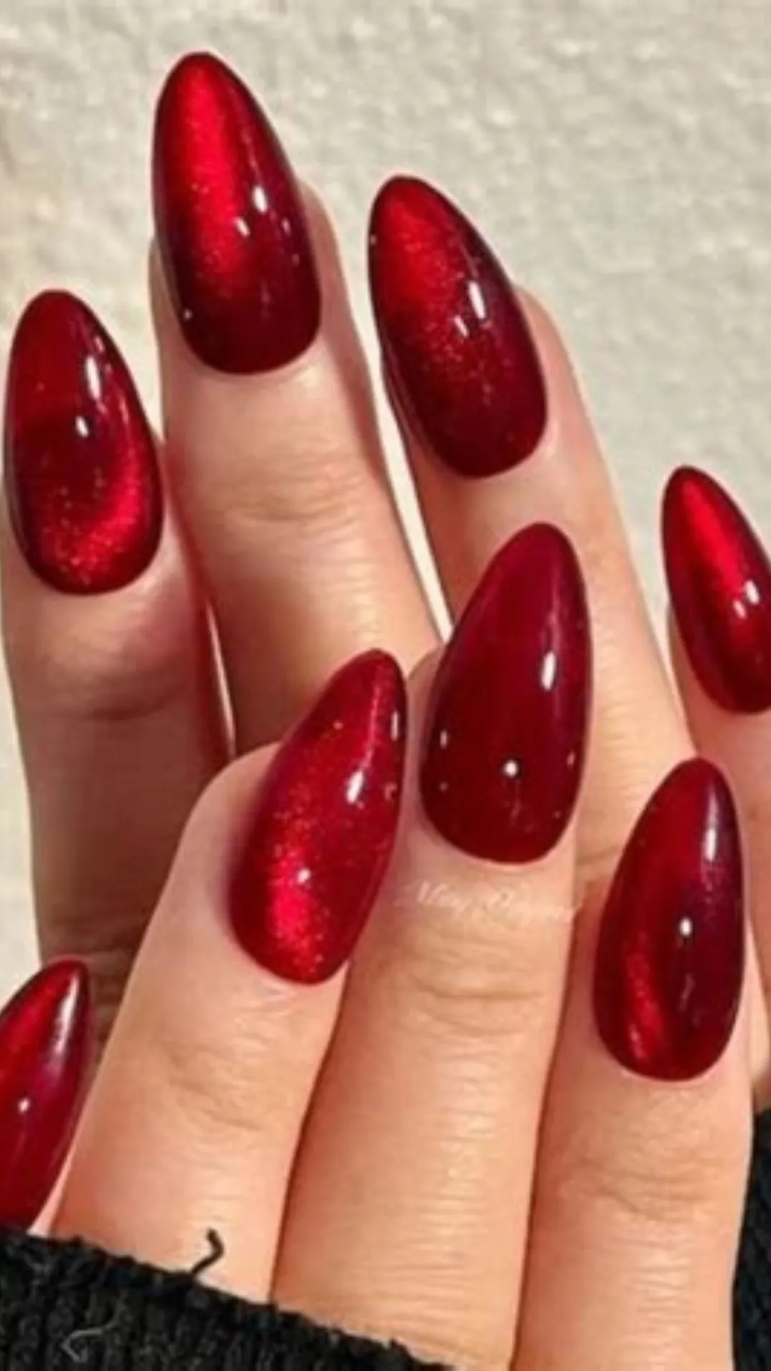 A close-up shot of hands displaying a captivating cat-eye velvet manicure on long, almond-shaped fingernails. The nail art uses a magnetic gel polish in a rich crimson or deep cherry red tone, creating a multi-dimensional, shimmering light effect that appears to glow from within as it catches different angles of lighting.