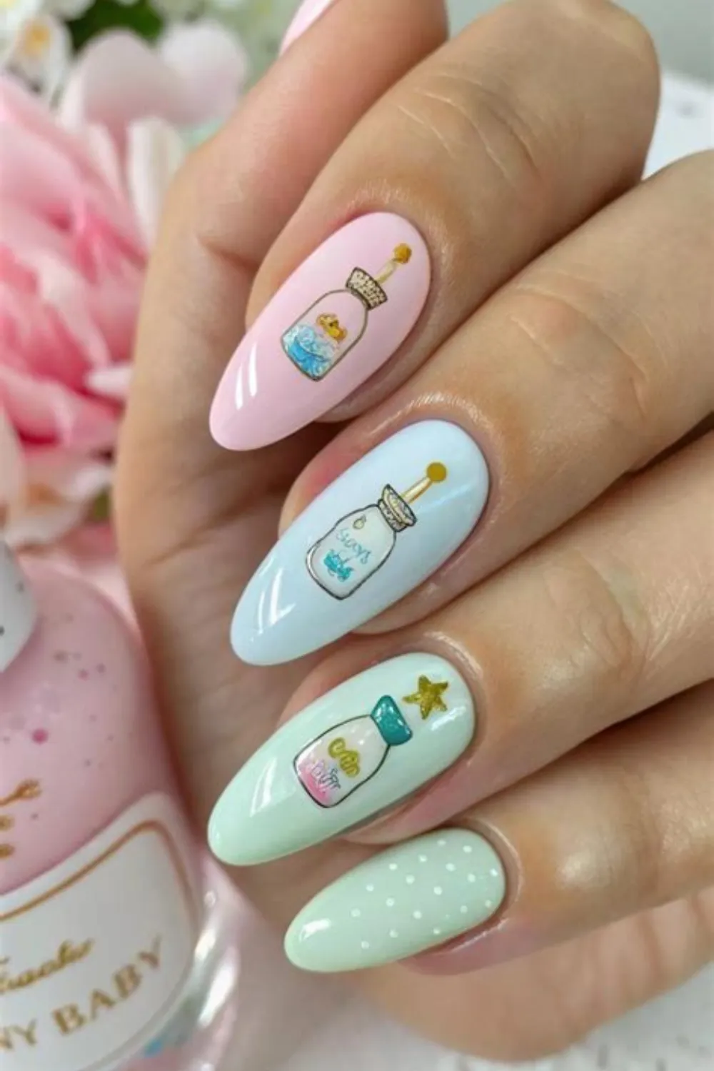A detailed close-up shot of a hand with long, almond-shaped nails showcasing a charming baby bottle theme. The nails alternate between soft pastel pink, powder blue, and mint green gel bases. Each nail bed features a delicate, vintage-style baby milk bottle illustration complete with a golden bottle cap, tiny star details, and soft pastel polka dot accent tips.