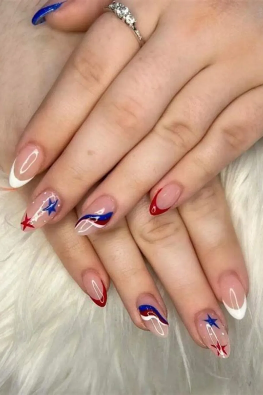 A close-up view of two hands resting on a fluffy white backdrop, displaying a chic patriotic French manicure on long, almond-shaped nails. The design features a mix of classic white French tips, asymmetric red and blue wave lines curving across sheer nude bases, and accent nails adorned with metallic red and blue stars alongside tiny white starburst dots.
