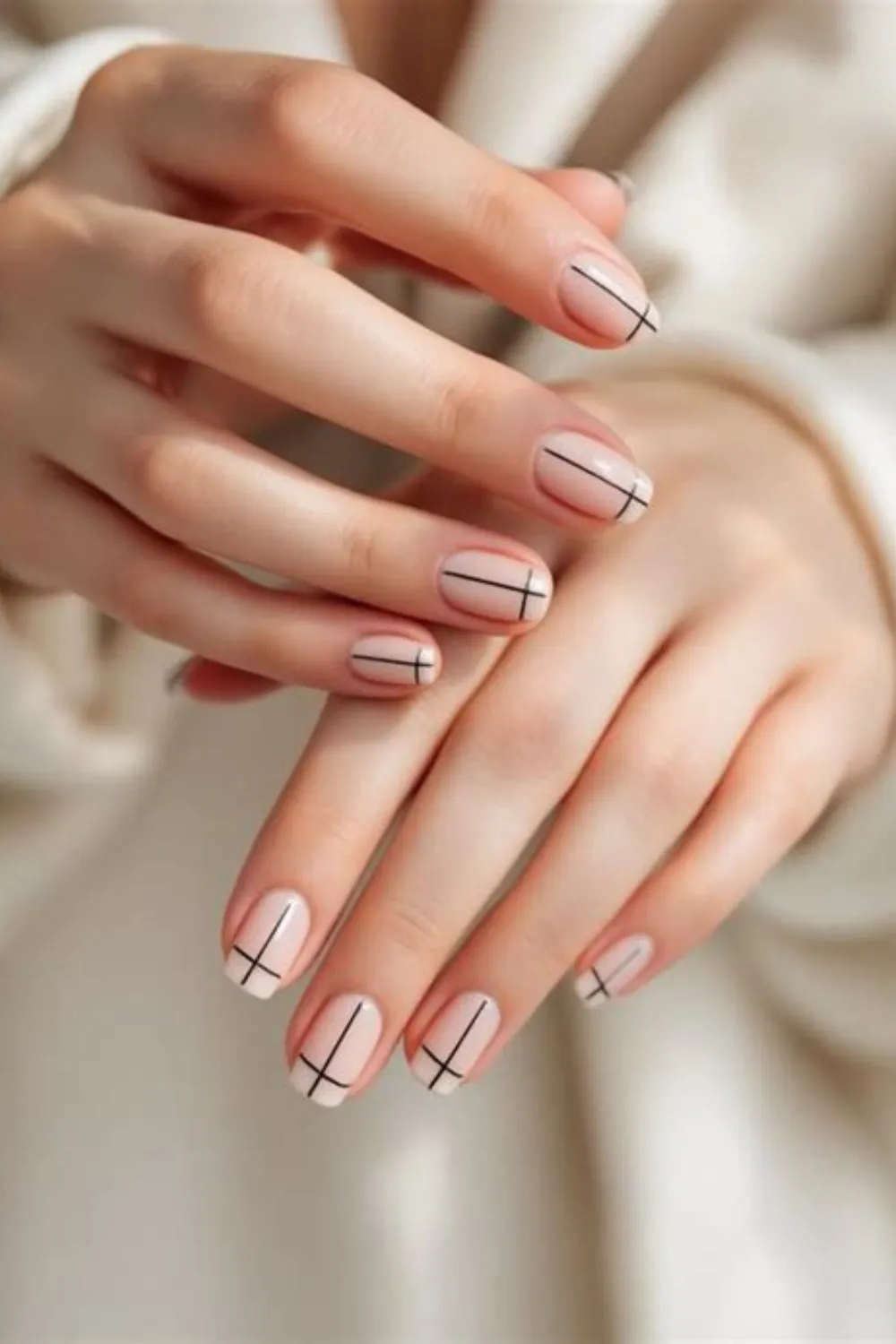 : A close-up shot of two hands displaying a sophisticated minimalist geometric manicure on short, oval-shaped nails. Over a smooth, semi-translucent nude base coat, fine black graphic lines intersect to form minimal cross or plus-sign patterns near the cuticles and side edges of each fingernail for an architectural look.