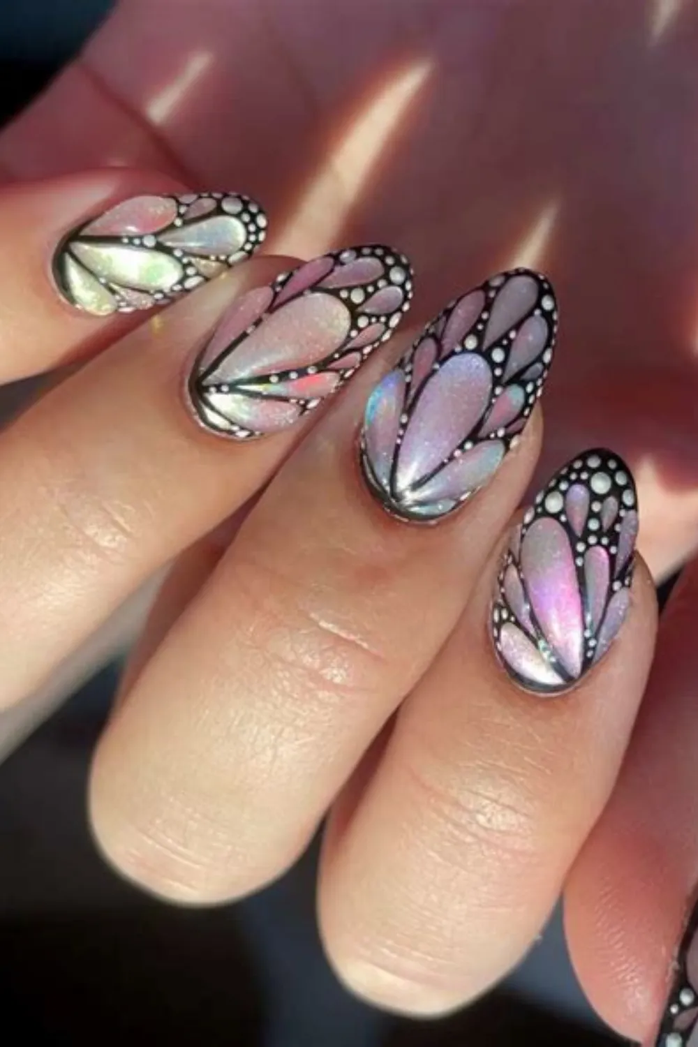 A close-up view of long almond-shaped nails featuring a dramatic, iridescent butterfly wing nail art design. The nails showcase an opal-like, shimmering pink and green cat-eye base coat framed by precise black outlines and fine white dots that realistically mimic the intricate texture and pattern of monarch butterfly wings.
