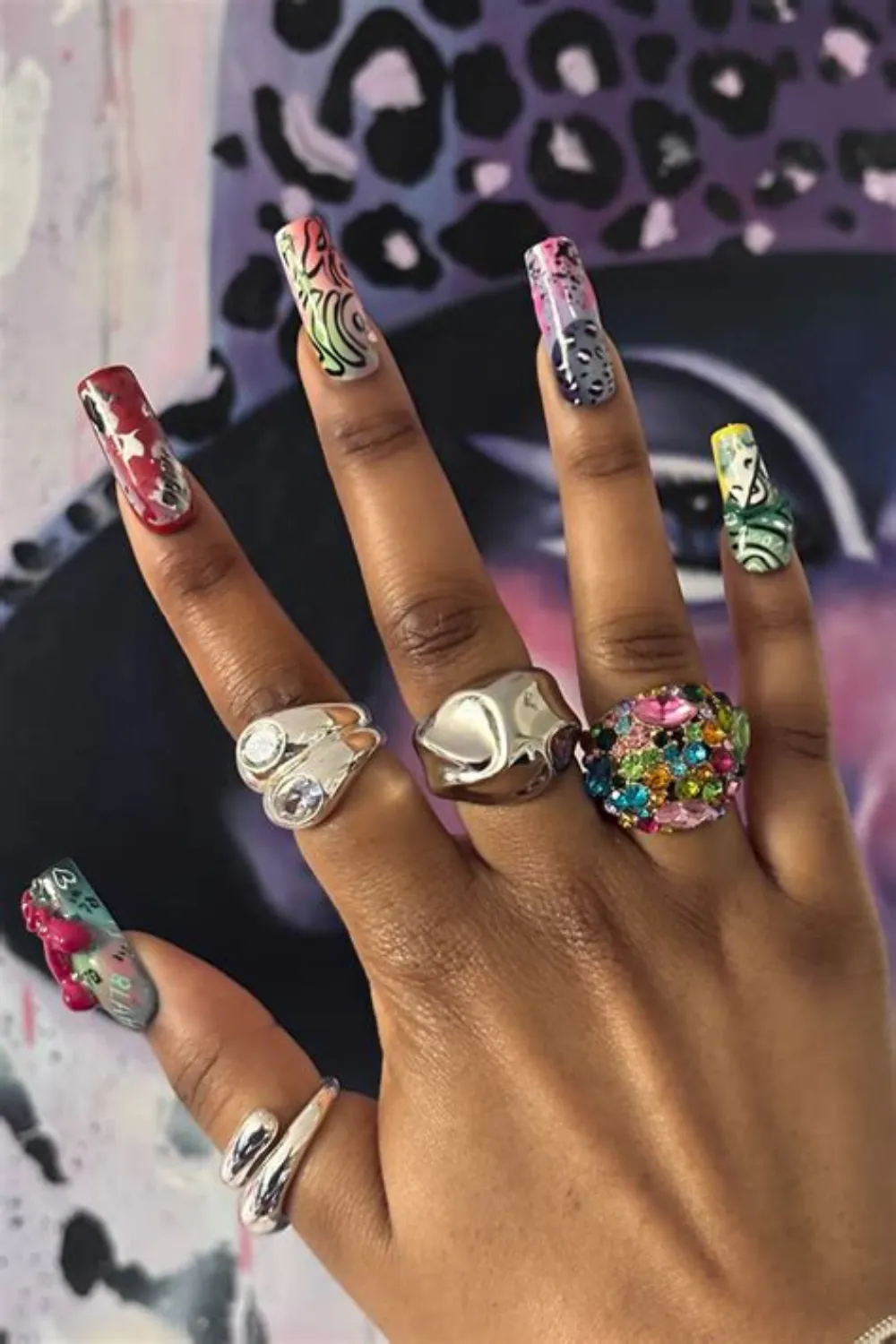 A close-up shot of a hand with long, coffin-shaped acrylic nails presenting an eclectic urban street-art design. Each nail displays a different chaotic abstract pattern blending neon graffiti, animal prints, and typography. The hand is heavily accessorized with statement chunky silver rings, a wobbly molten metallic band, and an oversized ring encrusted with multi-colored crystals.