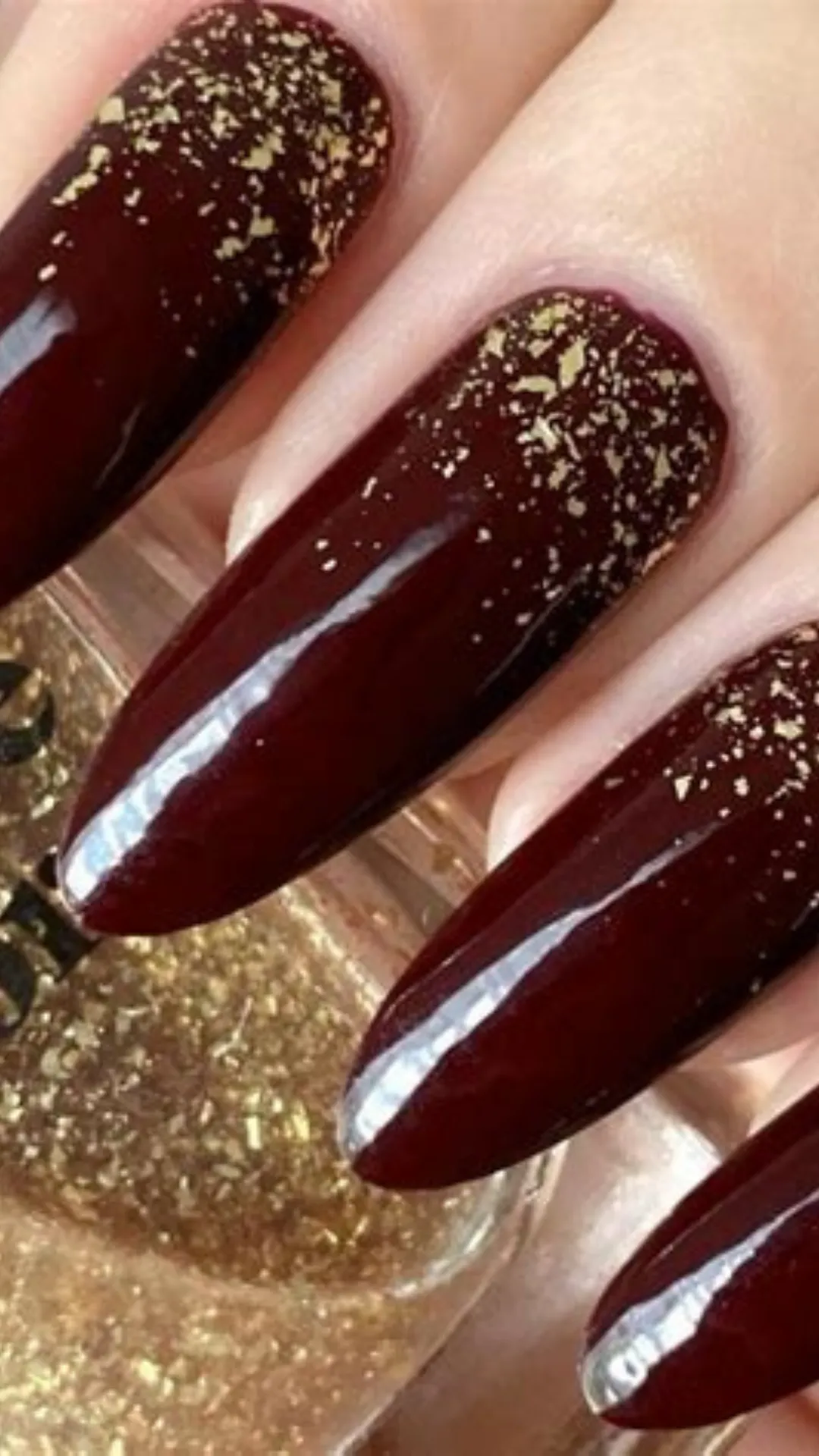 A luxurious close-up of long, glossy almond-shaped nails coated in a deep wine-red polish, accented by a delicate dusting of metallic gold foil flakes at the tips.