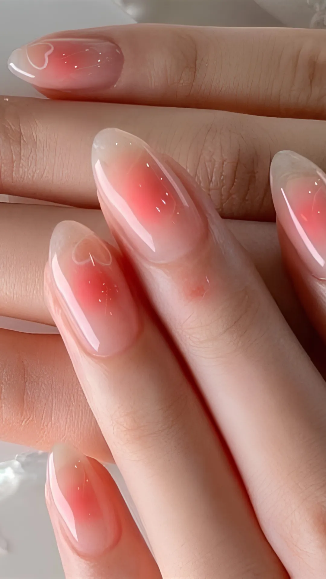 A detailed macro view of long, elegant oval-shaped nails showcasing a trendy Korean "blush nail" design. The nails have a clear, high-gloss base with a soft, airbrushed gradient of vibrant coral-red concentrated in the center of each nail bed. Delicate white hand-painted heart outlines and tiny faint sparkles sit elegantly over the blushed centers.