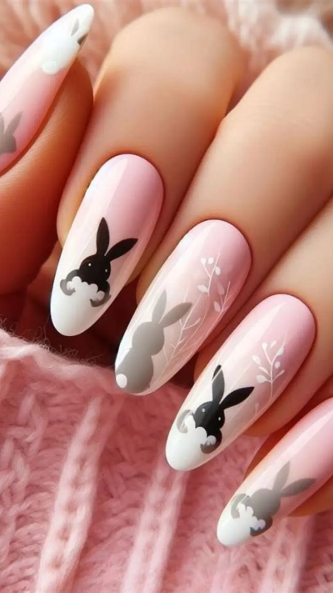 A close-up photograph of long, almond-shaped nails featuring a whimsical Easter theme. The manicure uses a glossy, semi-translucent light pink base that softly fades into white tips. Artfully painted across the nails are minimal silhouettes of grey and black Easter bunnies appearing to peek out from the edges, surrounded by delicate white botanical sprigs.
