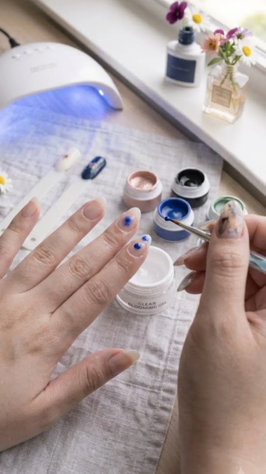 A step-by-step demonstration of blooming gel nail art, showing a detail brush applying small dots of royal blue gel polish onto a wet clear blooming gel base coat to create a bleeding watercolor flower petal effect.