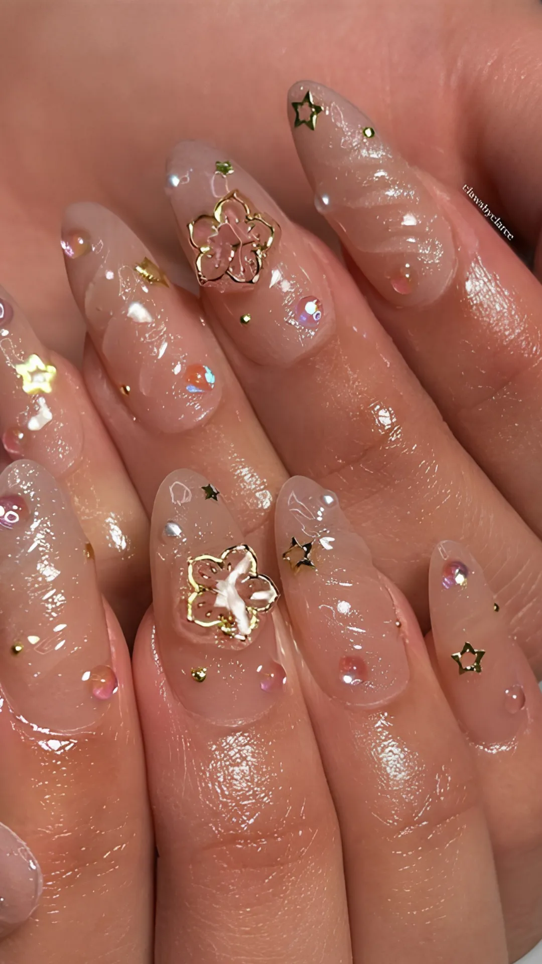 An extreme macro photograph of long, almond-shaped gel nails featuring a whimsical, clear-jelly fairycore design. The nails are covered in a glossy, translucent nude base embedded with multi-dimensional 3D elements. These decorations include small clear-sculpted flowers with golden metallic borders, reflective iridescent sequins, tiny gold star studs, and miniature faux pearls that give the manicure a wet, sparkling texture.