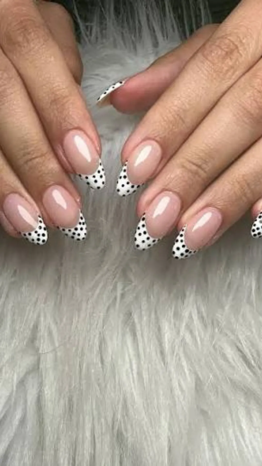 A photograph focusing on both hands displays a sophisticated, modern twist on the classic French manicure on almond-shaped nails. The nails feature a flawless, glossy nude base. In place of standard white tips, they showcase a clean white tip covered in precise black polka dots. The design is neat and minimalist while adding a touch of personality. The hands are posed gracefully against a soft, plush white faux fur background, emphasizing the elegant and clean lines of the manicured nails and fingers.