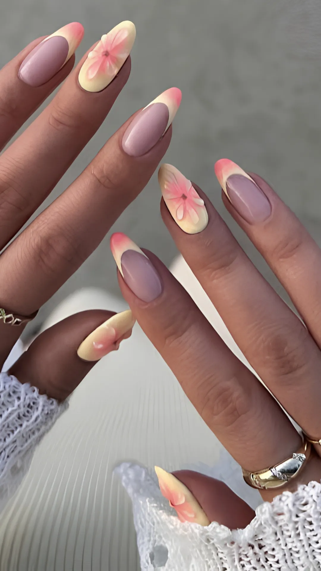 A detailed view of two hands showing long, stiletto-shaped nails with an elegant floral French manicure style. Most nails feature a muted mauve base with thin, curved pastel yellow French tips that fade into a soft pink hue at the absolute edges. The accent nails display a full pastel yellow surface decorated with a translucent 3D pink flower petal design that spans the center of the nail bed.