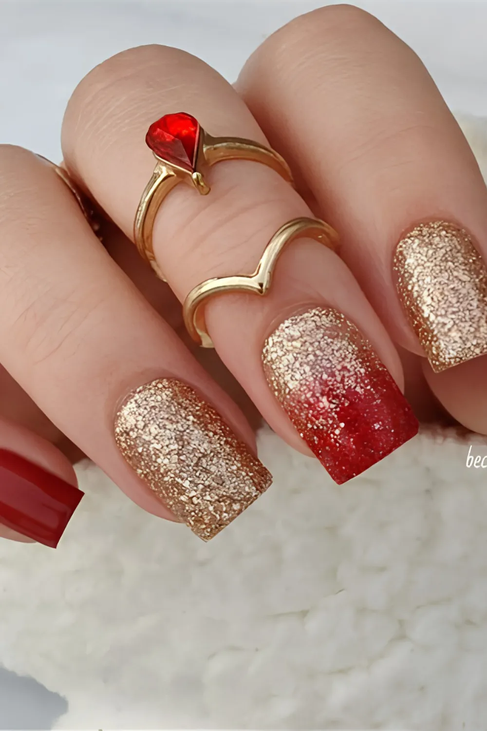 A glamorous close-up view of long, square-cut nails showcasing a high-contrast festive manicure. Several nails feature a dense, textured yellow-gold glitter lacquer, while the middle finger displays a seamless ombre gradient transitioning from the gold glitter at the cuticle to a rich ruby red at the tip. The model wears matching gold rings, including one set with a prominent red gemstone.
