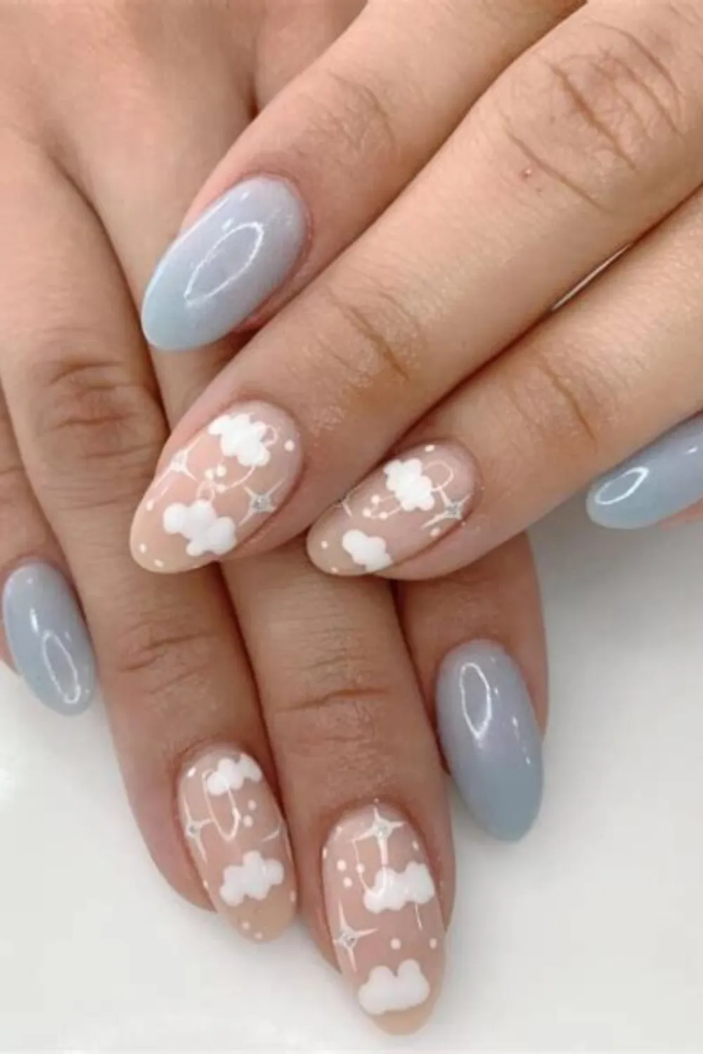 A close-up view of two hands displaying a dreamy, sky-inspired nursery manicure on almond-shaped nails. The index and pinky fingers are painted in a solid, high-gloss dusty blue polish. The middle and ring accent nails feature a sheer nude base decorated with fluffy white hand-painted clouds, delicate white starbursts, and tiny glittering rhinestones that sparkle under the light.