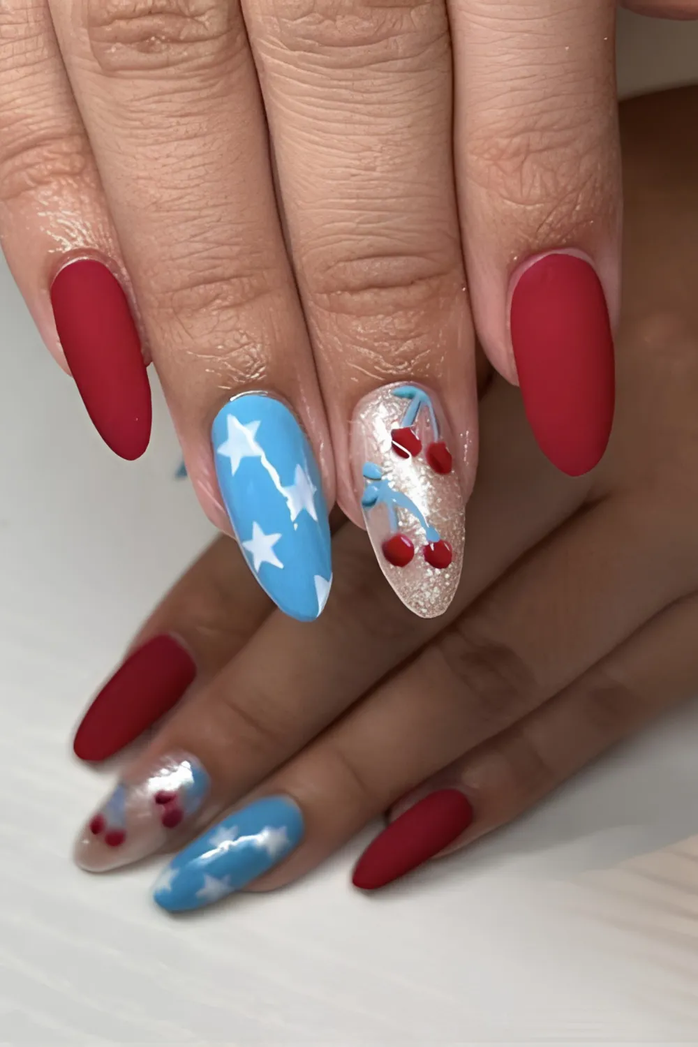 A detailed photograph of a hand with long, almond-shaped nails featuring a modern Fourth of July theme. The thumb, index, and pinky fingers are painted in a solid, velvety matte crimson-red polish. The middle finger is coated in a bright sky-blue lacquer with bold white stars, while the ring finger features a sparkling silver glitter base decorated with 3D cherry charms in red and blue.