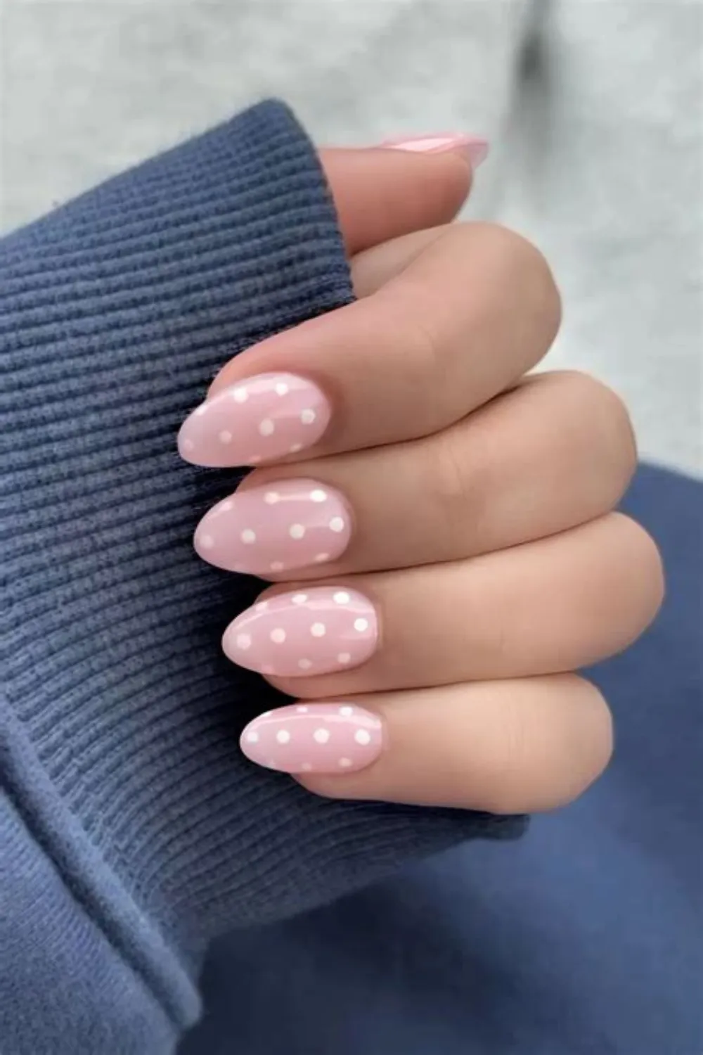A close-up view of a hand holding the ribbed cuff of a dusty blue sweater, showcasing a sweet polka dot manicure. The almond-shaped nails feature a soft, full-coverage pastel pink base coat decorated with evenly spaced, small white hand-painted polka dots across every nail for a clean, playful, and charming look.