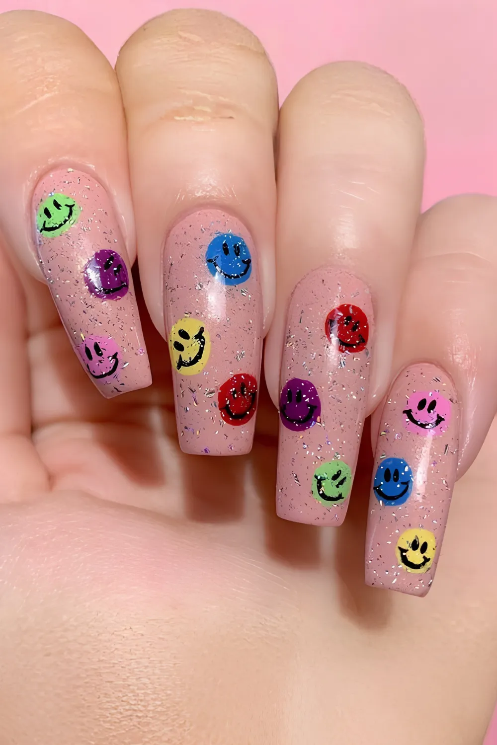 A detailed photograph of long coffin-shaped nails showcasing a retro smiley face theme over a dusty rose nude base coat filled with fine silver foil flecks. Scattered across each nail bed are multi-colored hand-painted smiley faces in neon green, bright blue, purple, yellow, and red, bringing a playful 2000s pop-art vibe.