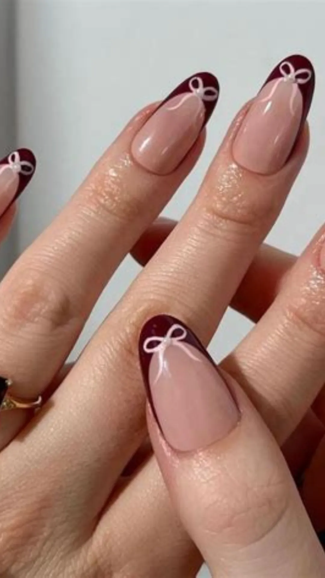 Coquette-style holiday nail art featuring a sheer nude base with sophisticated burgundy French tips, each decorated with a delicate white painted bow.