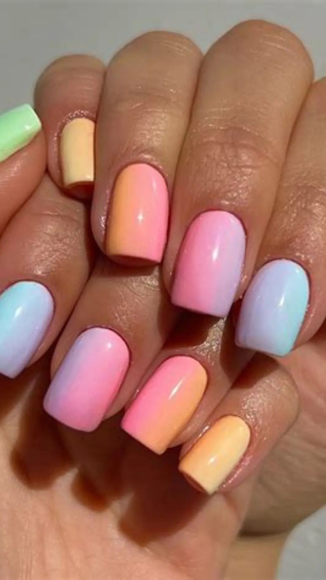 A vibrant showcase of a glossy, multi-colored pastel gradient manicure on short, squared nails. The nails feature smooth, seamlessly blended vertical ombré transitions between classic spring colors, including soft sky blue, lavender purple, bubblegum pink, warm peach, and muted yellow. The colors blend beautifully across both hands for a bright, cheerful aesthetic.