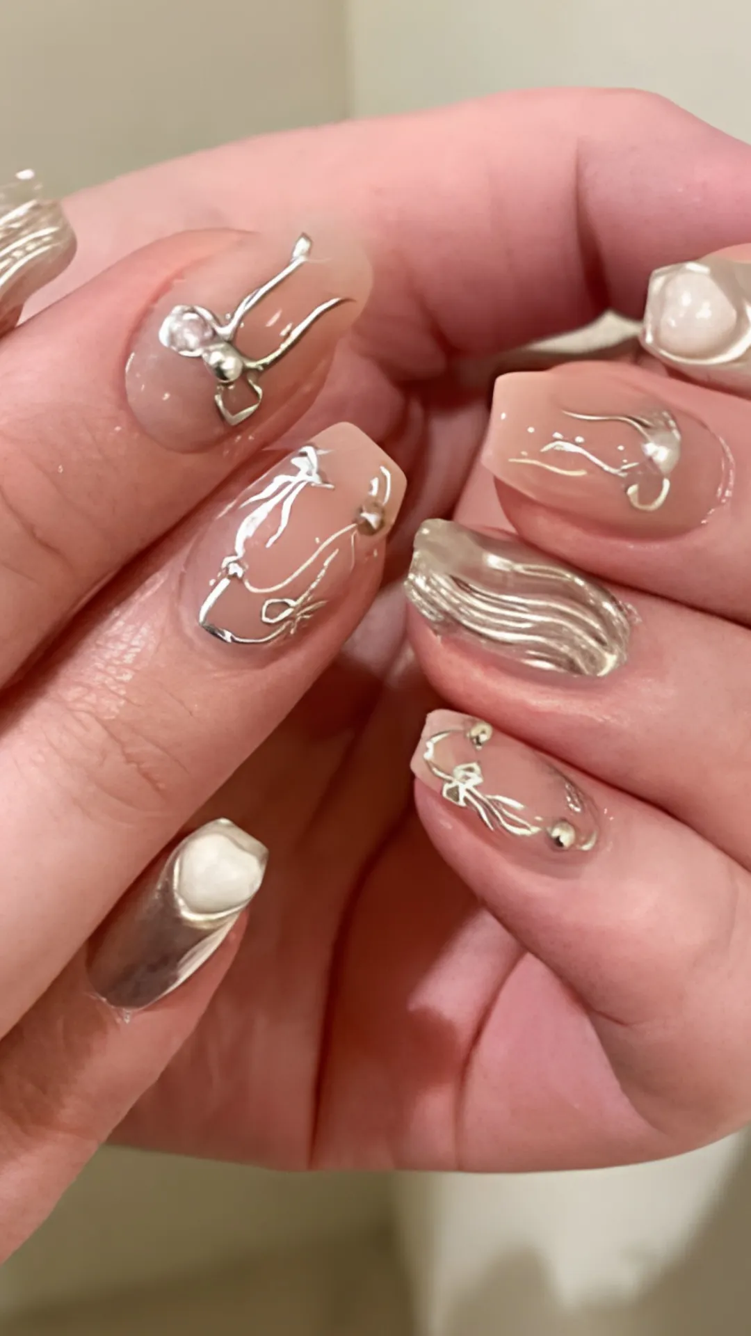 A close-up shot of a pair of hands displaying a modern, edgy Y2K-inspired gel manicure on medium-length coffin nails. Over a smooth, semi-sheer nude gel base coat, the nails are decorated with abstract, liquid-like 3D chrome squiggles and sculpted metallic silver bows. One accent finger stands out with a fully textured, molten silver chrome finish, while the thumb features a chunky 3D white heart charm.