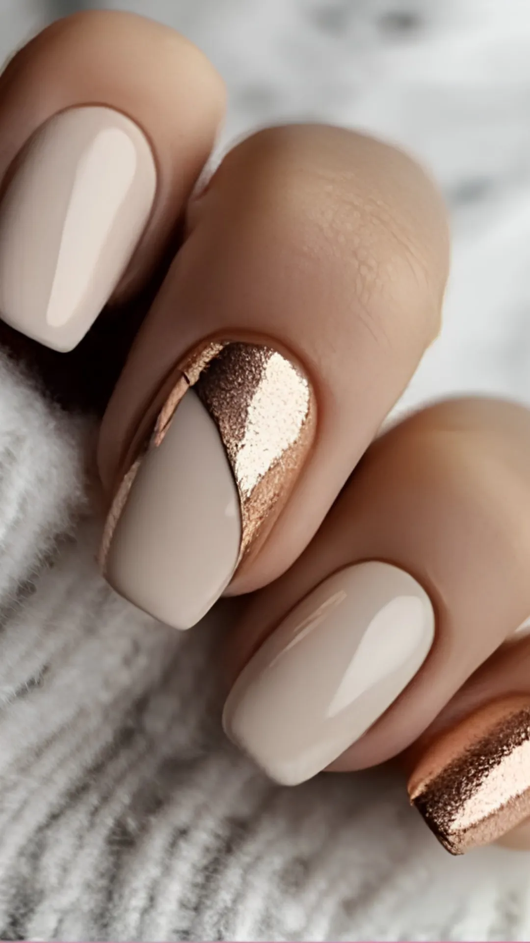 An extreme close-up of a flawless, high-contrast chic manicure on square-shaped nails. The index and ring fingers are painted with a smooth, full-coverage glossy cream-beige lacquer. The middle finger features an asymmetric color-blocked pattern, dividing the cream-beige polish from a textured, metallic rose gold foil sections that gleams under the light. The hand rests against a white textured fabric.