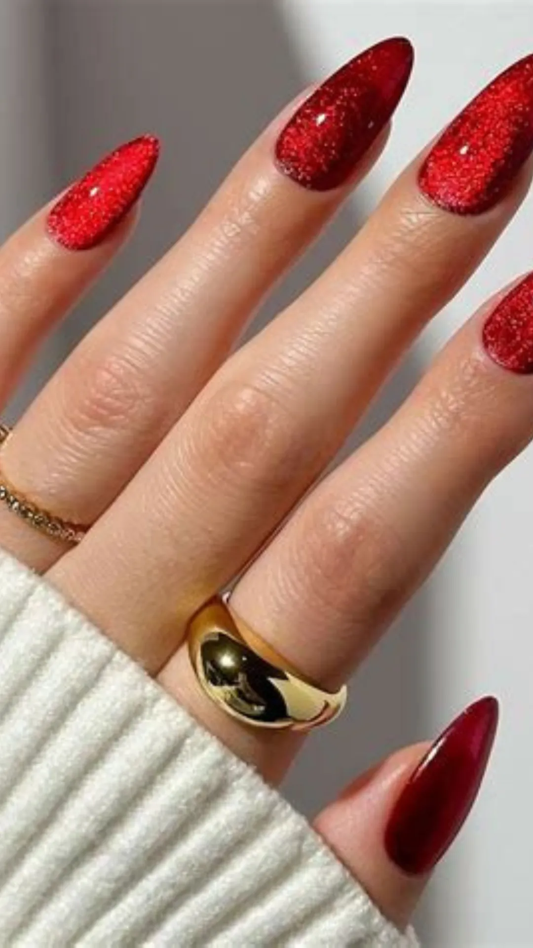A detailed photograph of a hand showcasing a sparkling texture-rich manicure on long stiletto nails. The nails are coated in a dense, dazzling ruby red glitter lacquer that catches the light with thousands of tiny reflections. The hand is accessorized with a thick, chunky polished gold dome ring on the ring finger and a thin gold band on the pinky finger.