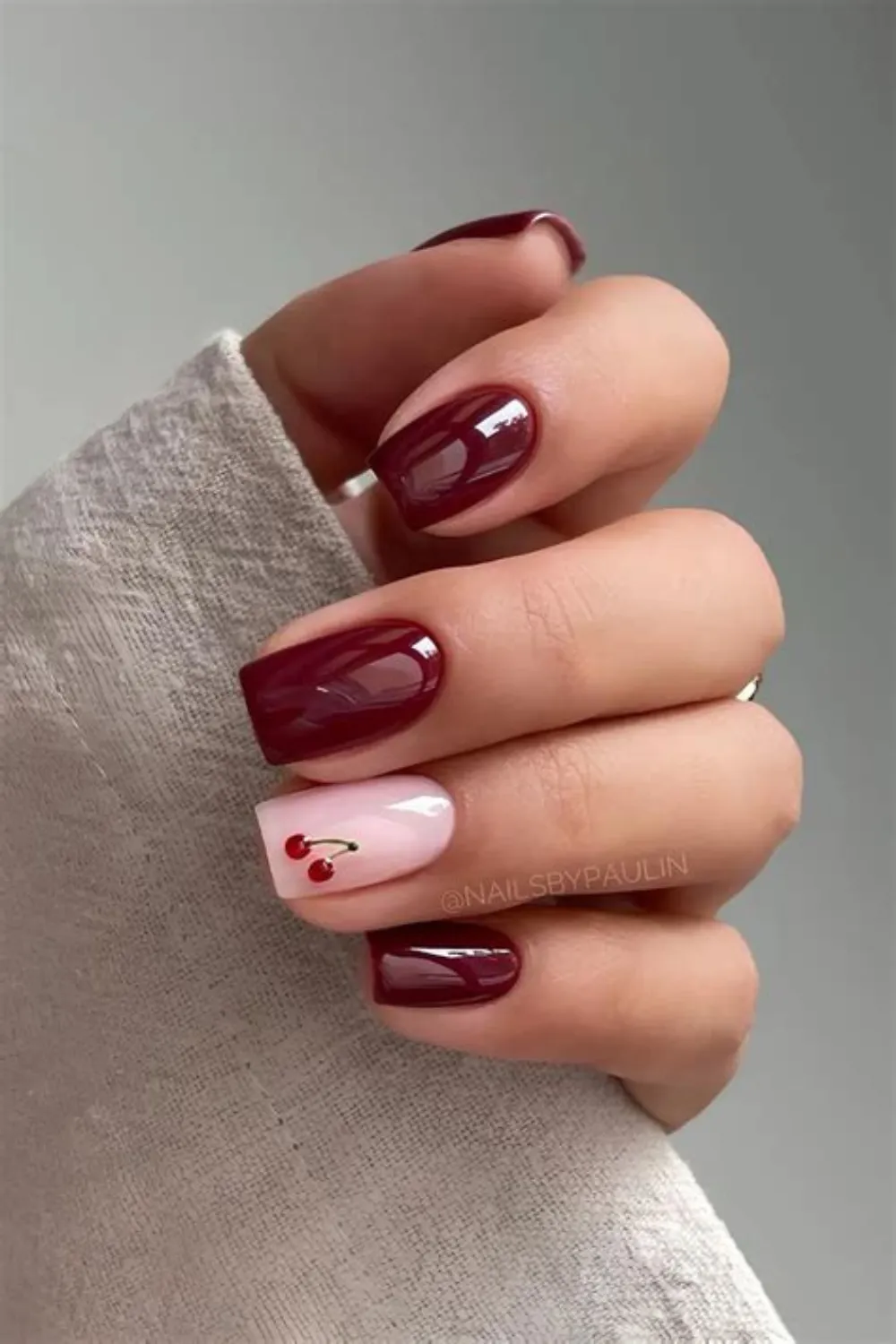 A photograph focusing on a sophisticated mix-and-match manicure on short, squared nails. The thumb, index, middle, and pinky fingers are painted with a dark, ultra-glossy oxblood red polish. The ring finger serves as a delicate accent nail, featuring a milky white translucent base decorated with a tiny, hand-painted pair of red cherries on a green stem.