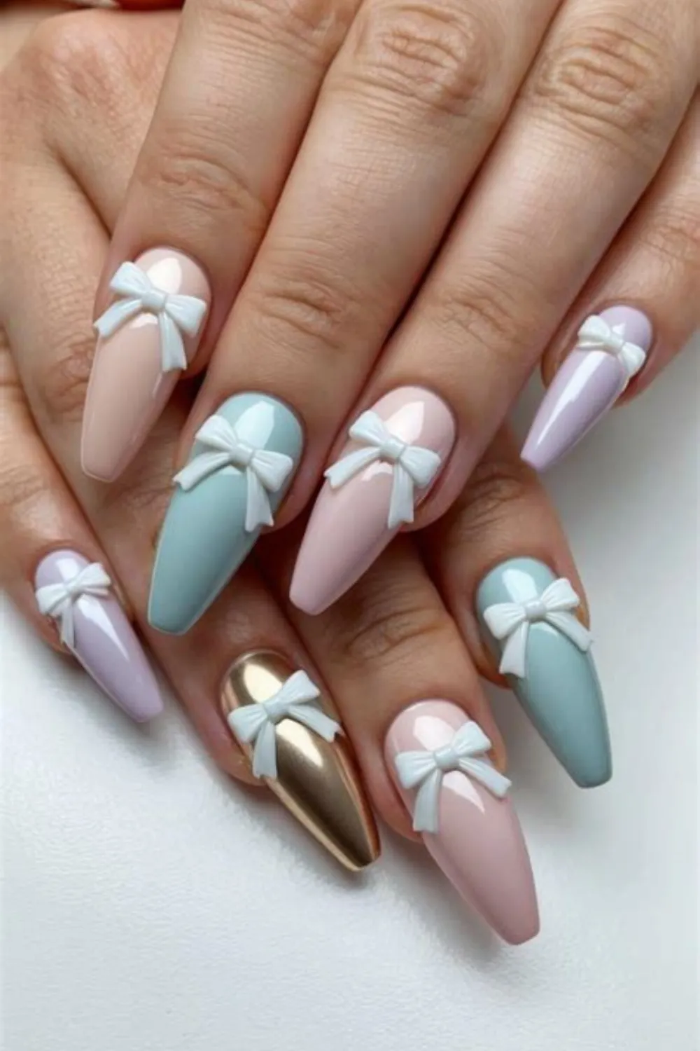 A detailed photograph showcasing a luxurious 3D bow manicure on long, coffin-shaped nails. The design mixes delicate pastel shades, including baby pink, soft lilac, muted teal, and a shiny metallic gold accent nail. At the center of every single nail bed rests an oversized, sculpted white acrylic bow charm that gives the manicure a playful, gift-wrapped aesthetic.