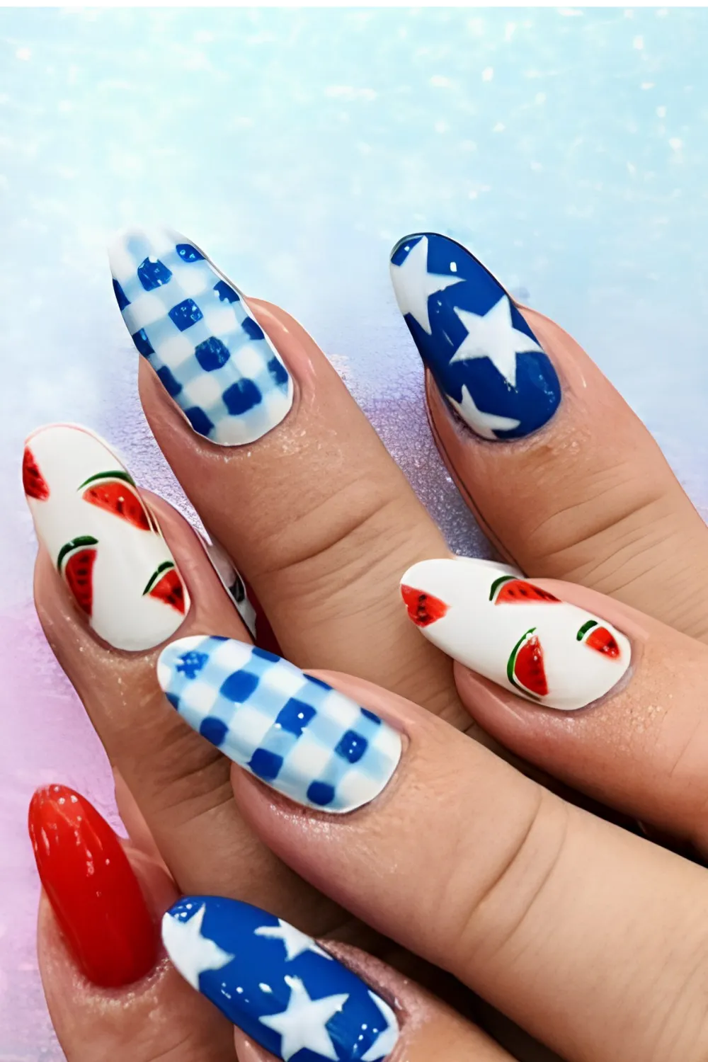 A close-up shot of a summer picnic and 4th of July inspired manicure on long oval nails. The design incorporates an eclectic mix of patterns across fingers, including a classic blue-and-white gingham check pattern, solid royal blue nails with large white stars, a bright red accent nail, and crisp white nails decorated with painted red watermelon slices.