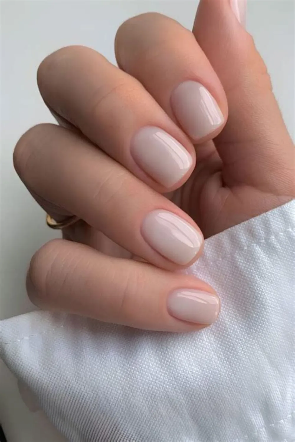 A close-up photograph focusing on an ultra-clean, monochromatic nude gel manicure on short, rounded fingernails. The nails are painted in a smooth, high-gloss ivory-beige shade that closely matches the skin tone, creating a flawless "soap nail" appearance against the crisp white fabric of a shirt cuff.