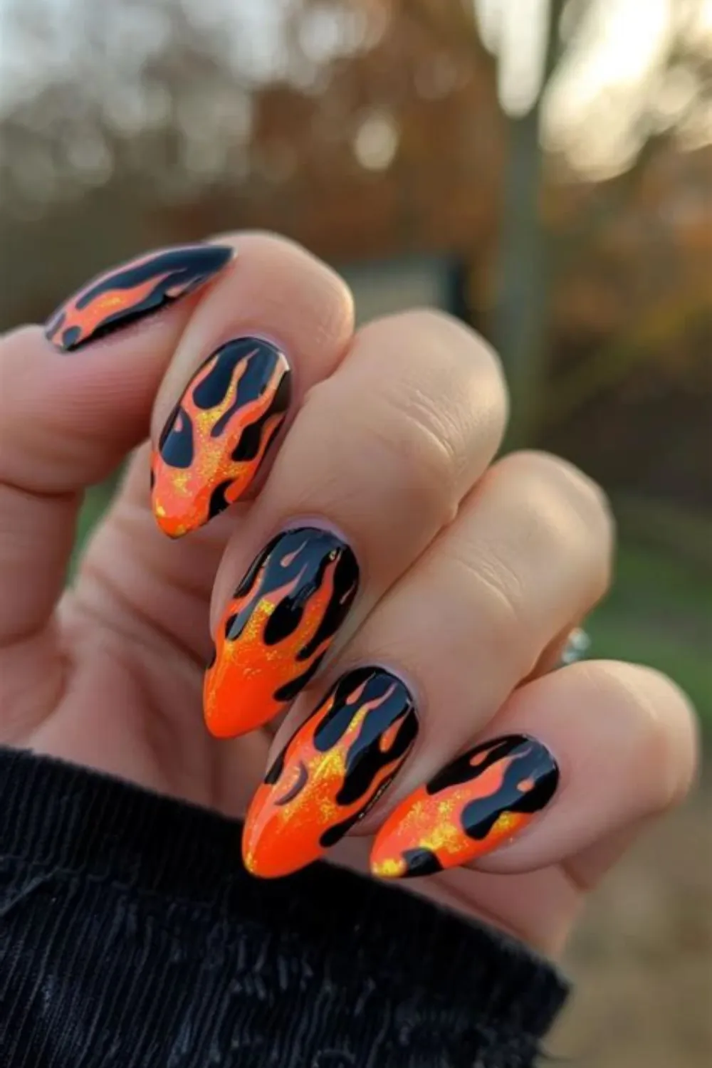 A close-up photograph of a hand held outdoors displaying long, almond-shaped flame nail art. The nails feature a vibrant neon orange and yellow glitter base that transitions into glossy black flames licking upward from the cuticles and sidewalls, creating a bold hot-rod aesthetic iconic to late 90s and early 2000s streetwear fashion.
